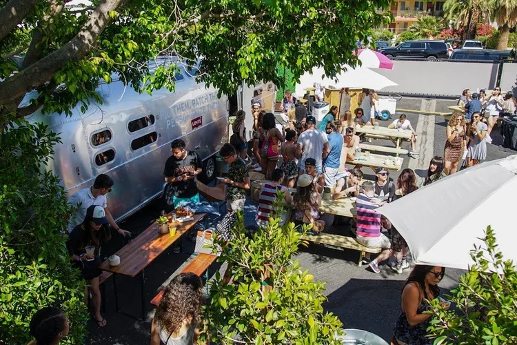 People gathered outdoors around picnic tables and a food truck on a sunny day. Some people are standing and some are sitting under umbrellas. There are trees and cars in the background.