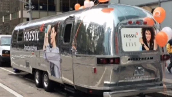A shiny, vintage-style trailer with advertisements for Fossil, a fashion accessories brand, featuring a woman with dark curly hair on the side and back of the trailer.