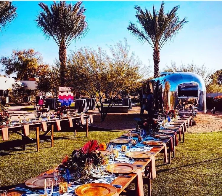 Outdoor banquet setup with long tables decorated with flowers, plates, and glasses, palm trees in the background, and a shiny silver food truck.