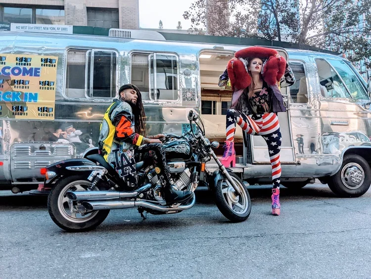 A woman poses with one leg elevated on a motorcycle in front of a vintage silver Airstream trailer, with a man sitting on the motorcycle and a city street background.