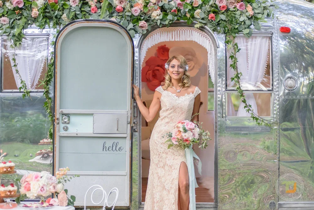 A bride in an elegant lace wedding dress holding a bouquet of pink and white flowers, standing at the entrance of a decorated vintage trailer adorned with pink roses and greenery, with white curtains and a sign that says 'hello'.