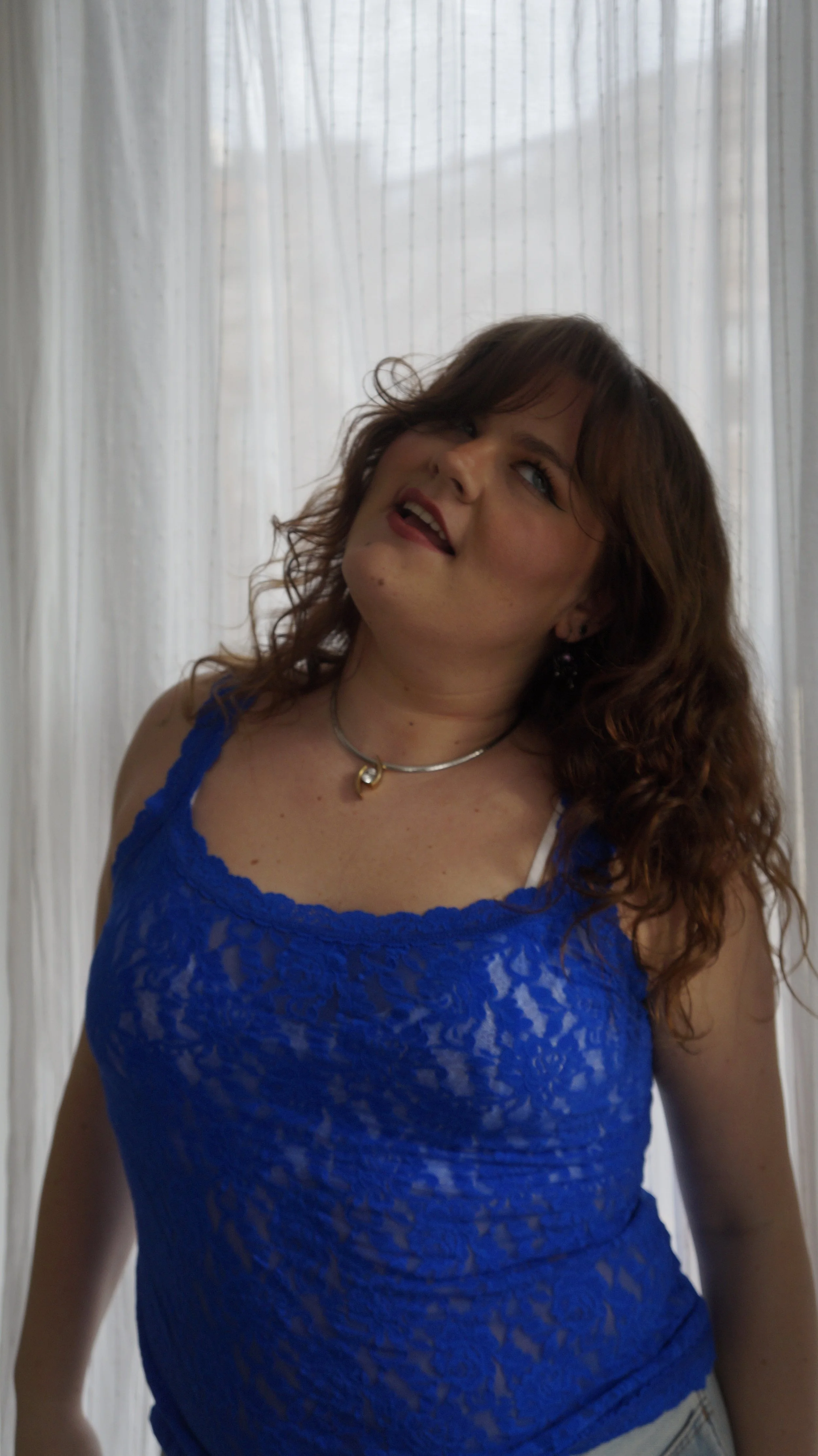 A woman with wavy brown hair wearing a blue lace top, a silver necklace, standing in front of a sheer white curtain.