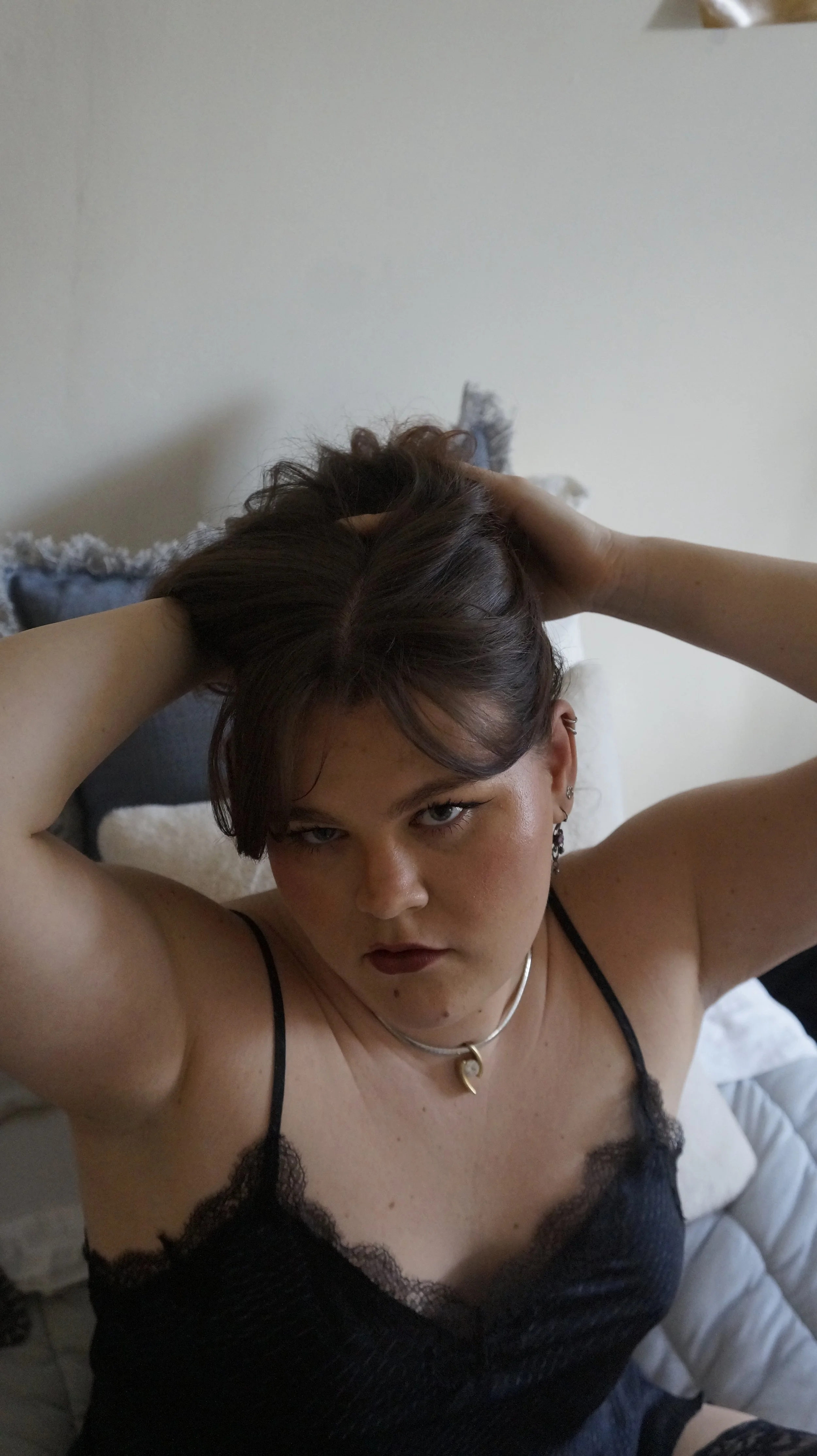 A person with dark hair and makeup wearing a black lace camisole, holding their hair up with both hands, standing in a room with a white wall and a bed with pillows in the background.