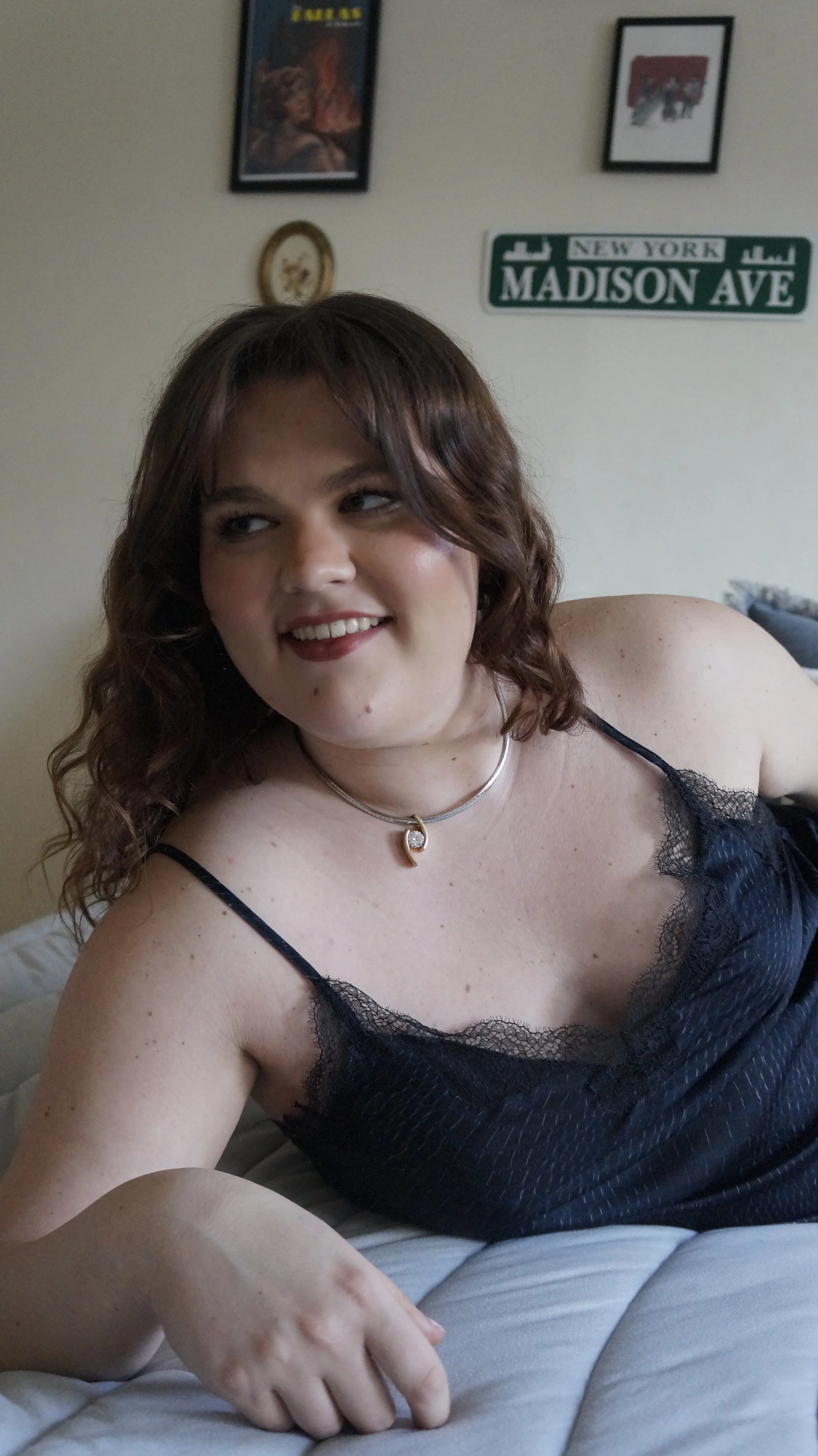 A young woman with curly brown hair, wearing a black lace-trimmed camisole, lying on a bed, smiling and looking to the side. The background features a wall with framed art and a street sign that reads 'Madison Ave' in New York.