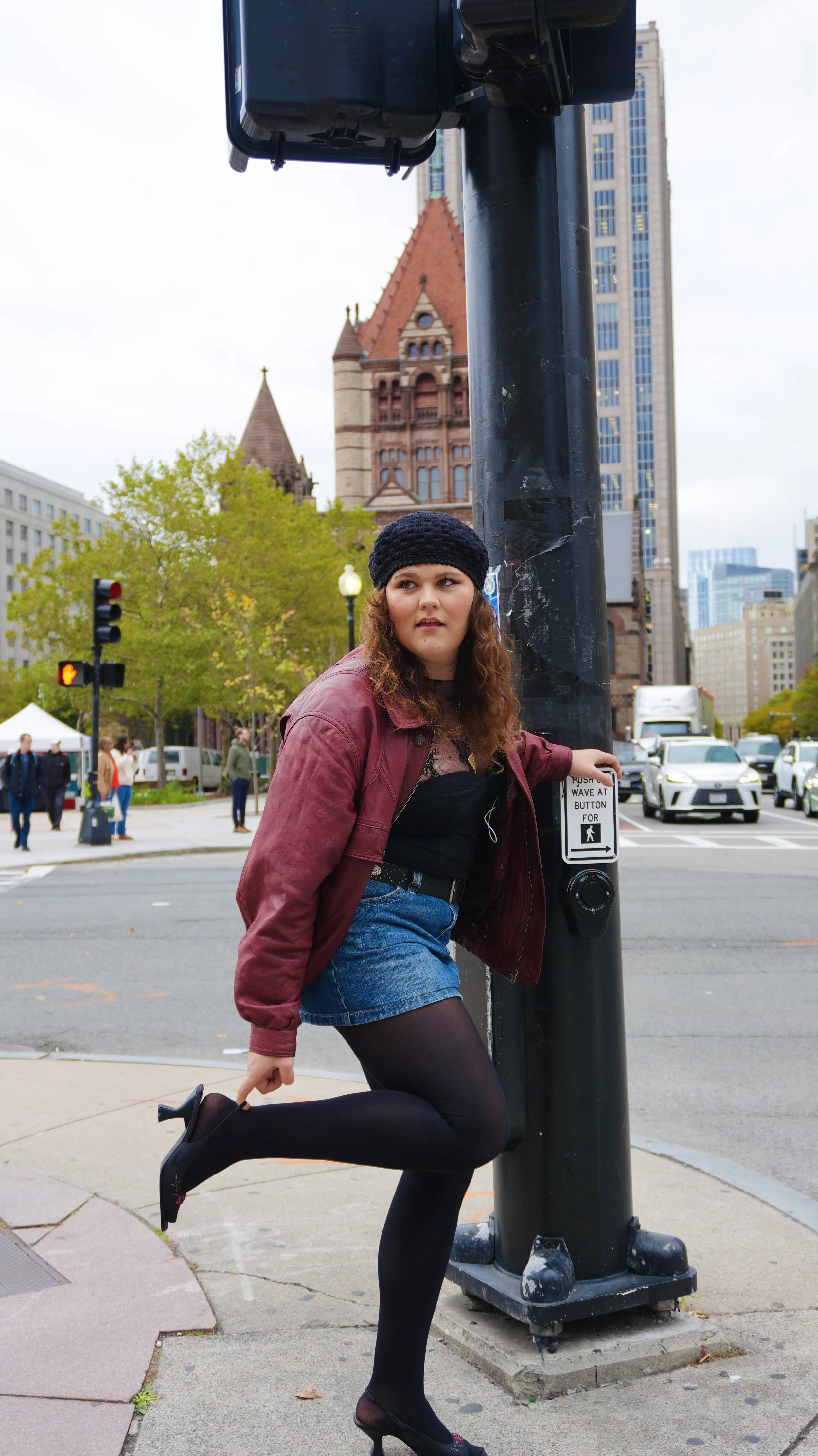 A young woman in black tights, a denim skirt, black top, maroon jacket, and black knit hat stands at a crosswalk button on a city street, with a historic building in the background.