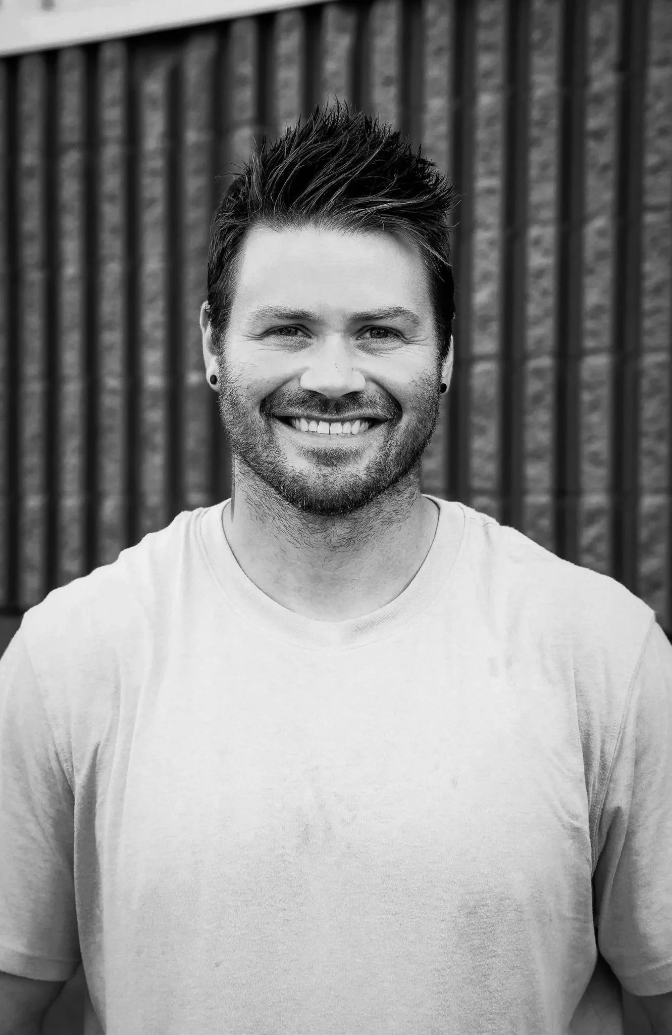 Dalton Nowicki, owner of Reformed Painting Co, wearing a light-colored T-shirt, standing outdoors in front of a textured fence background.