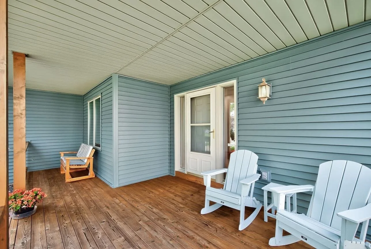 Covered porch with wooden floor, two white Adirondack chairs, a small side table, a wooden bench with cushions, a flower pot, and a blue house exterior with windows, a door, and a wall-mounted light fixture.