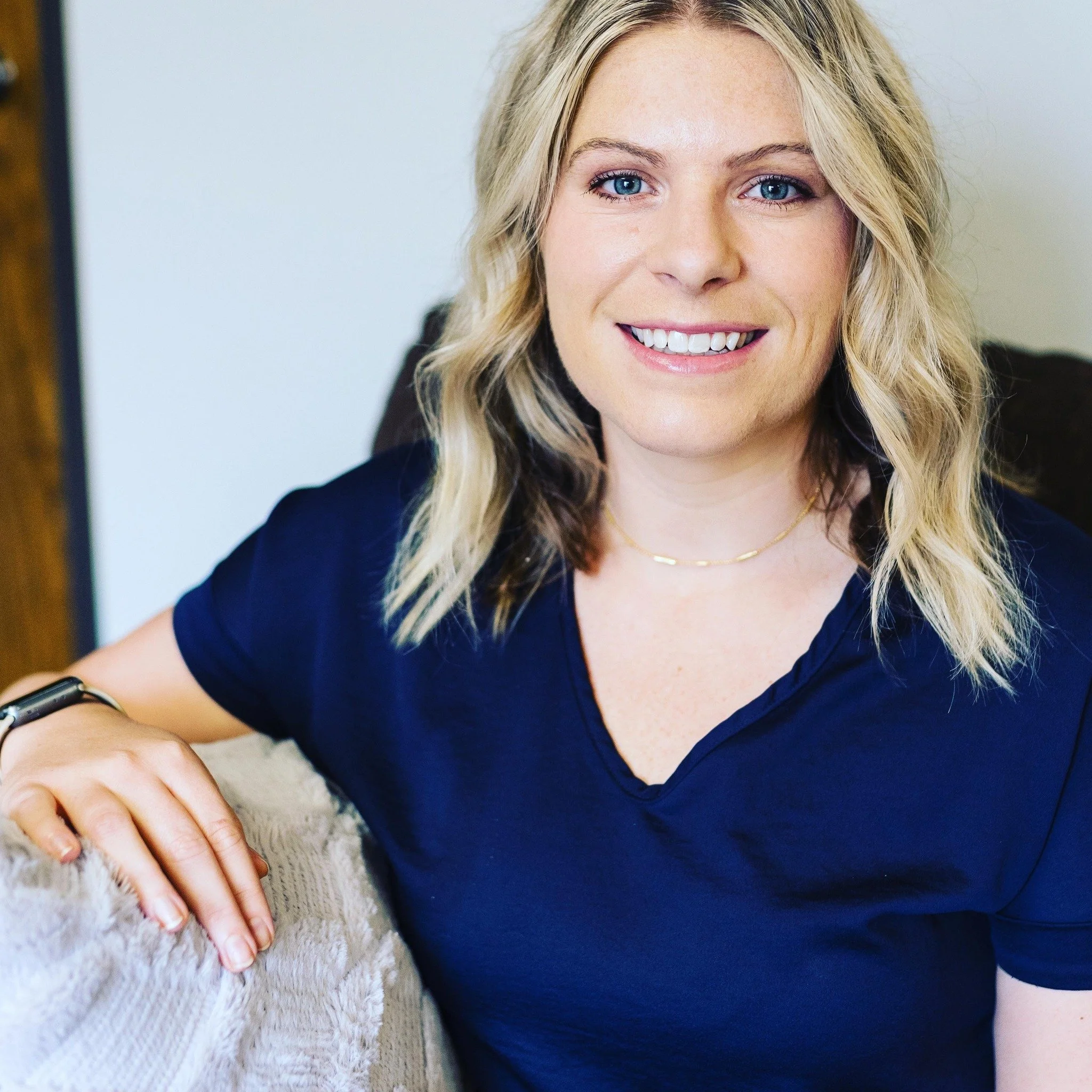 A smiling woman with blonde hair, blue eyes, wearing a navy blue top, a delicate gold necklace, and a black watch, seated on a beige sofa.