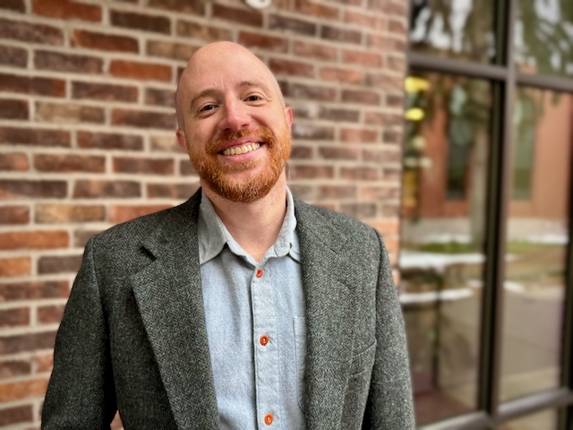 Man with a beard smiling, standing outdoors in front of a brick wall and window.