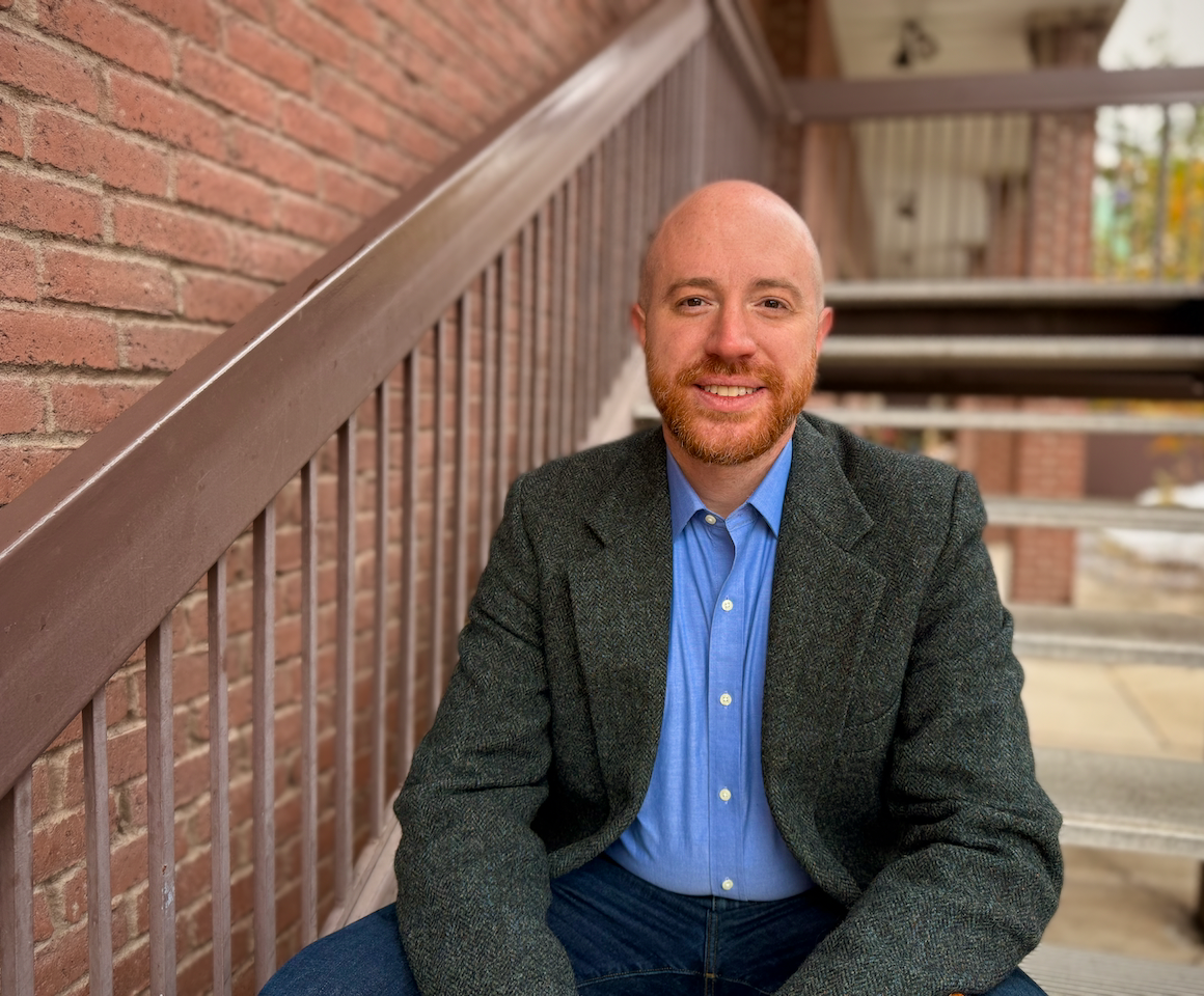 A smiling man with a beard and bald head sitting on outdoor stairs, wearing a dark blazer and light blue shirt, with a brick wall behind him.