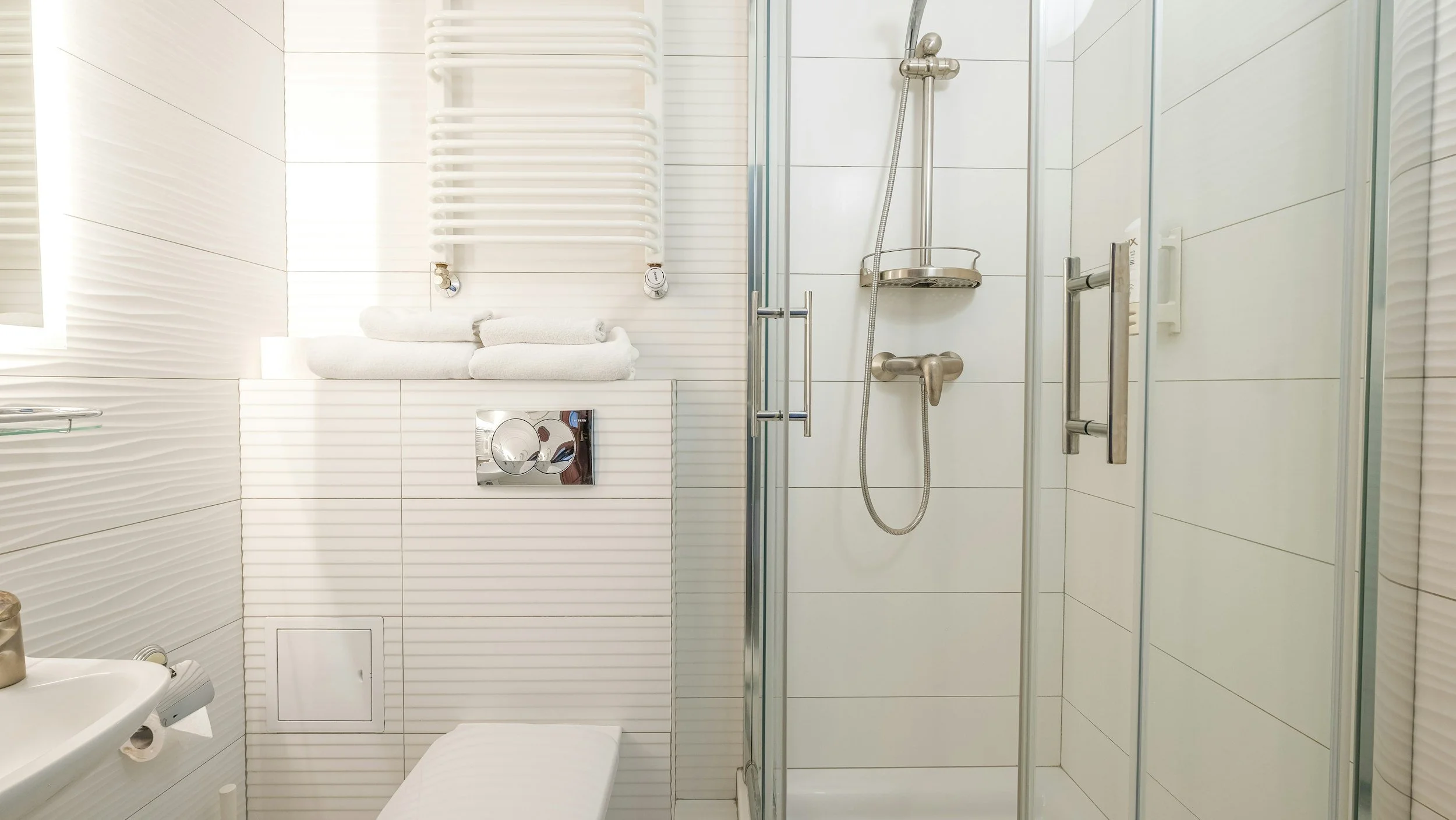 Small modern bathroom with white tiled walls, a glass shower enclosure, a wall-mounted toilet, a towel rack with folded towels, and a small window.