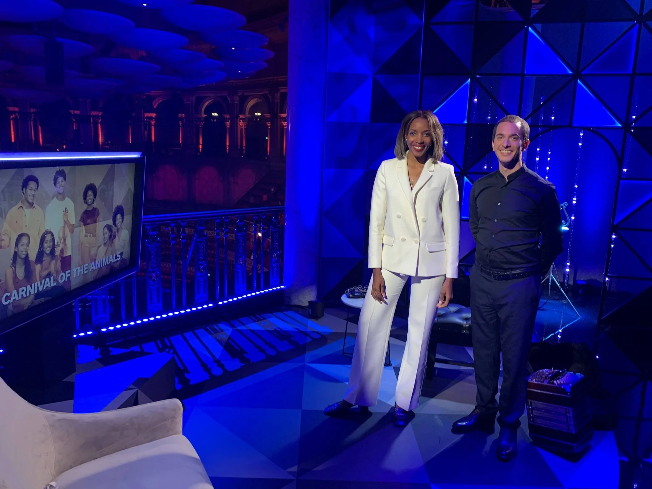 A TV interview set of BBC 4 in the Royal Albert Hall at the BBC Proms in 2022 with Josie d'Arby and Guillermo Willis, bandoneon player, sitting across from each other in a dark, modern studio with blue and purple lighting and geometric decor.