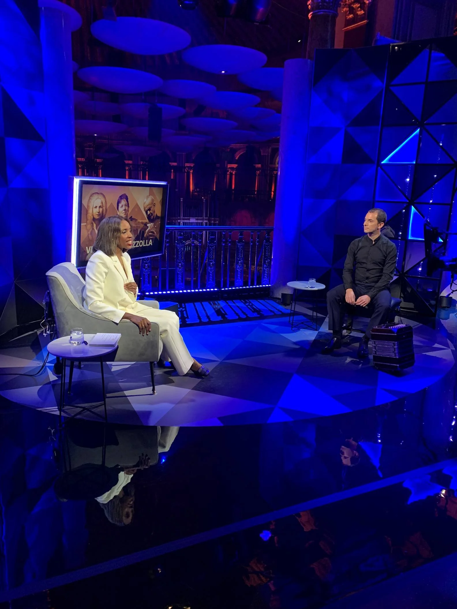 A TV interview set of BBC 4 in the Royal Albert Hall at the BBC Proms in 2022 with Josie d'Arby and Guillermo Willis, bandoneon player, sitting across from each other in a dark, modern studio with blue and purple lighting and geometric decor.