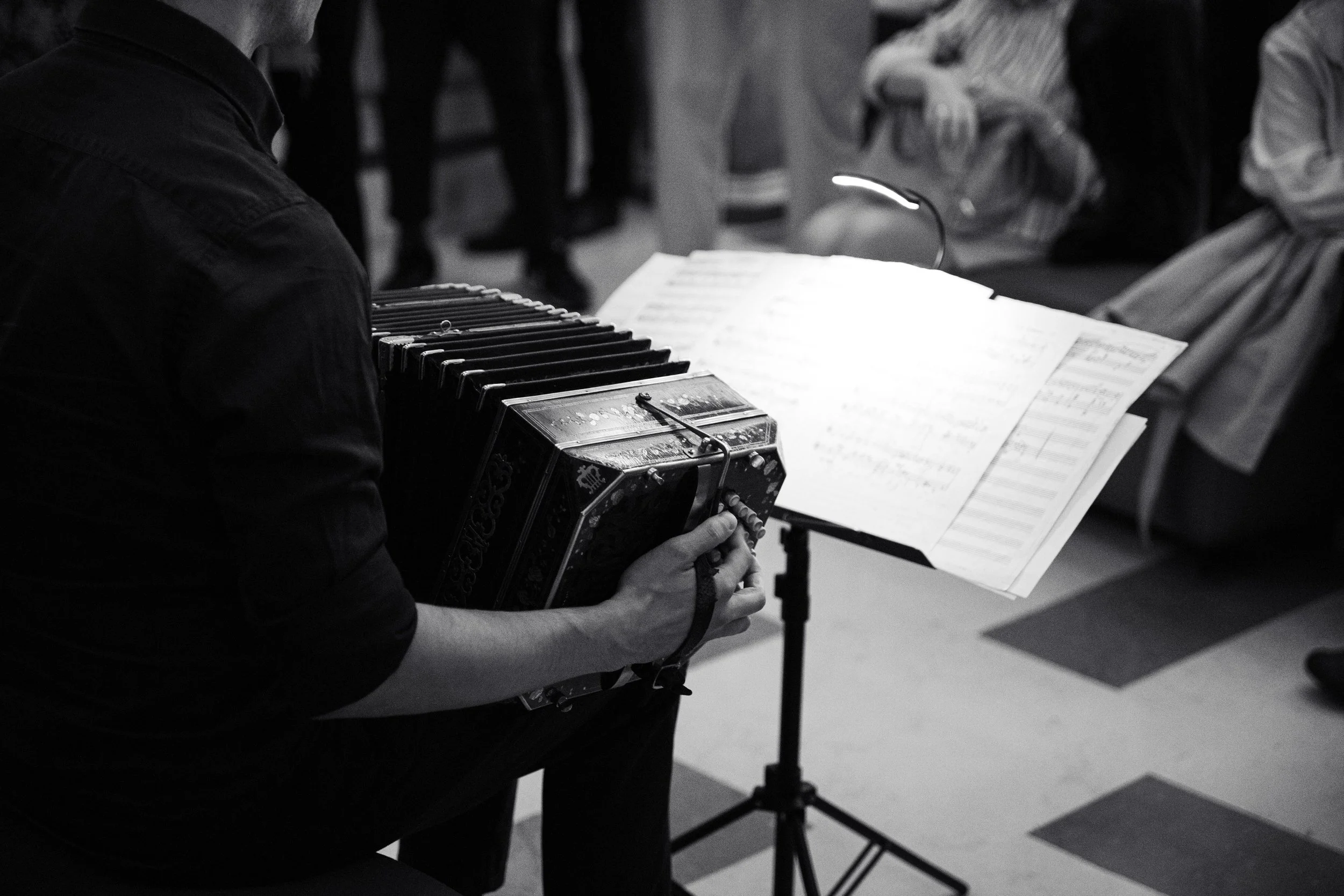 Guillermo Willis playing Tango in a bandoneon in a room, with a music stand holding sheet music, and people sitting in the background.