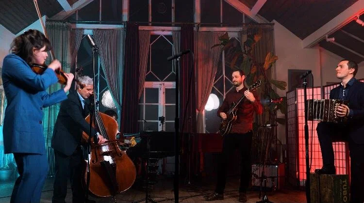 Desbande Quintet in 2023 performing Tango of Astor Piazzolla on a stage with musical instruments, including a bandoneon, violin, a double bass, and two guitars, in an indoor venue with colorful lighting.