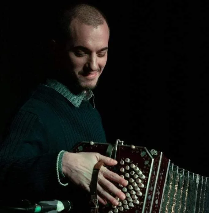 Photo of Guillermo Willis playing Tango music with a bandoneon, smiling against a black background.