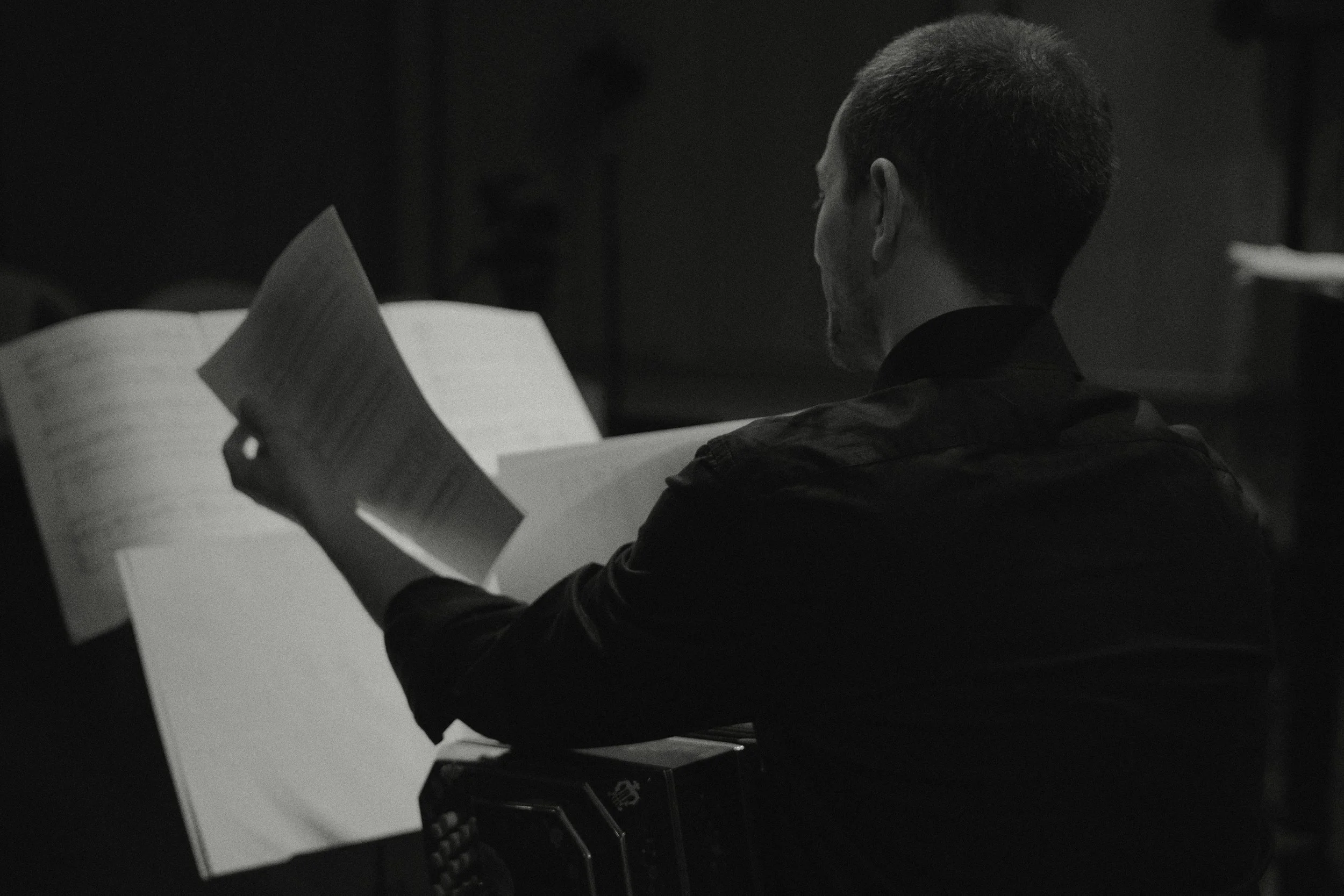 Black and white photo of Guillermo Willis playing Tango music with a bandoneon, viewed from behind, reading sheet music in a dark room.
