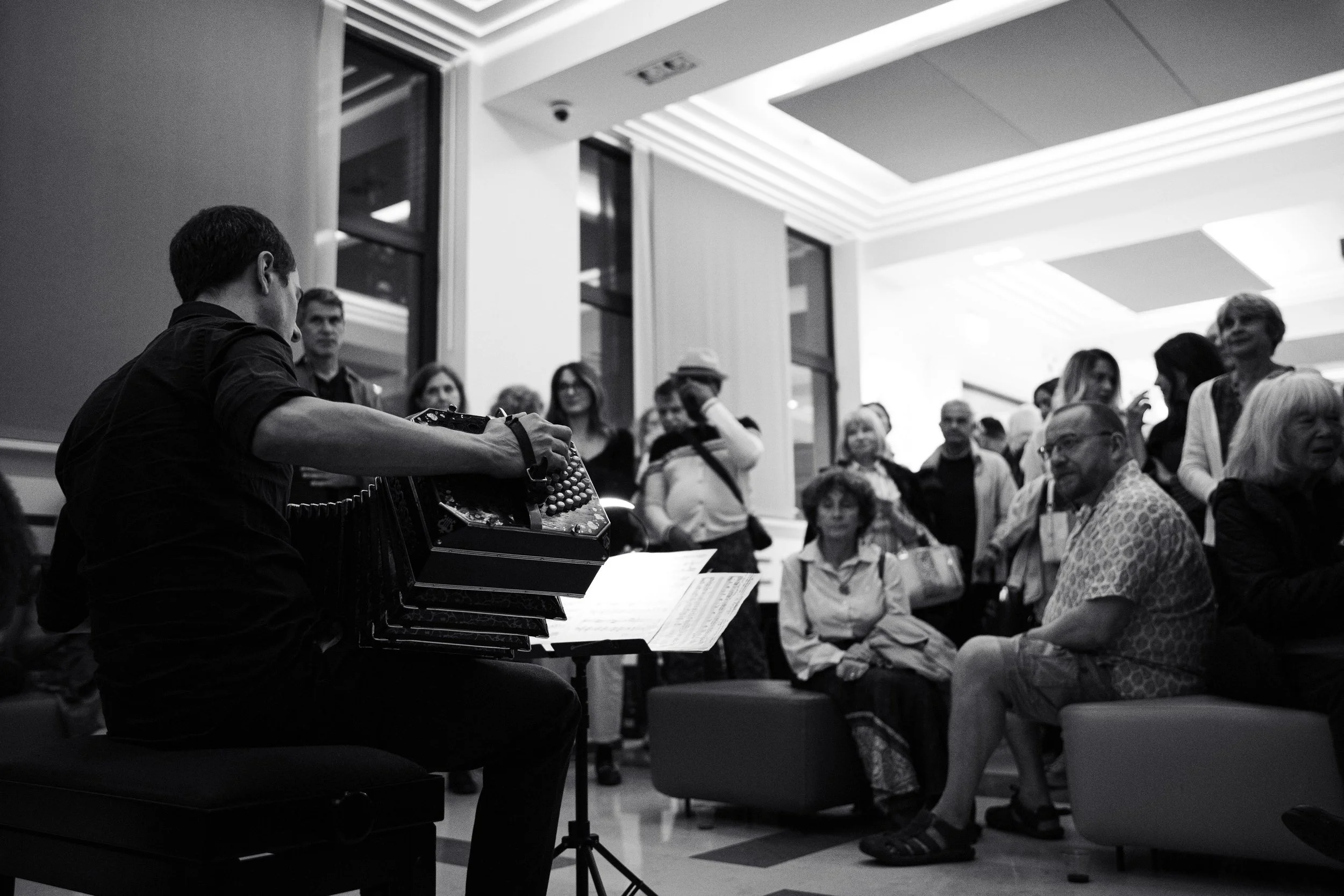 Black and white photo of Guillermo Willis playing Tango music with a bandoneon in front of a group of people indoors. The audience is seated and standing, watching the performance.