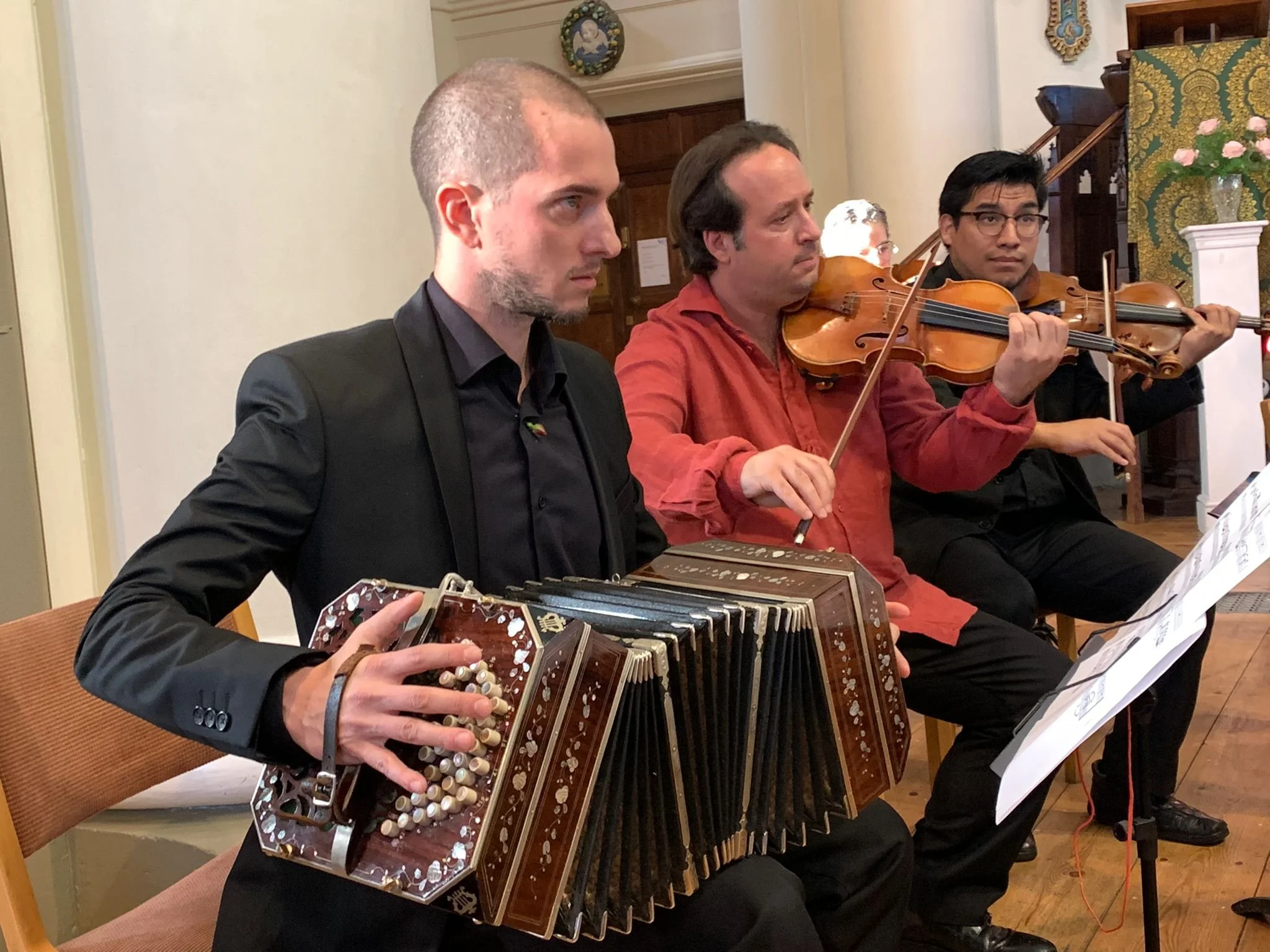 Three musicians performing together: Guillermo Willis playing Tango music with a bandoneon, a man playing a violin, and another man playing a violin, in an indoor setting with decorative elements in the background.