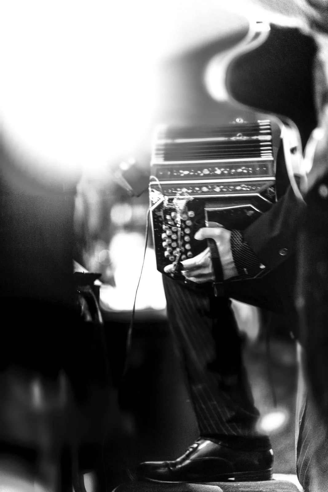 Guillermo Willis playing Tango in a bandoneon in black and white, wearing a dark suit with pinstripes, standing on stage with a spotlight shining from behind.