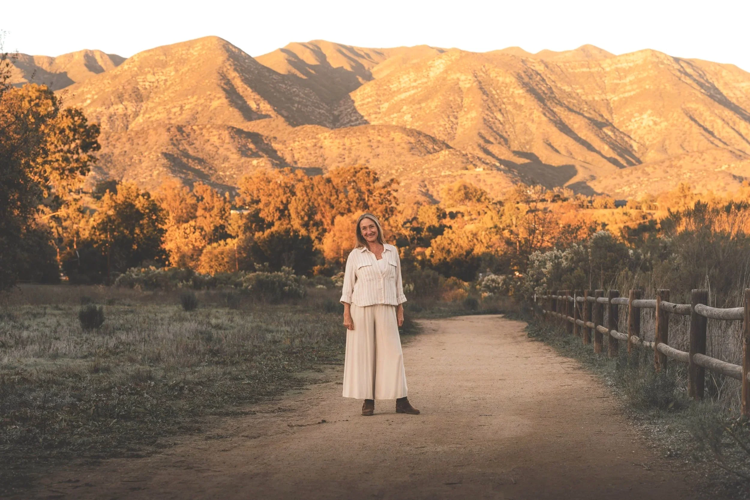 Amyris Wilson stands on a dirt path in Ojai, California, with the Topa Topa mountains and trees illuminated by a golden sunset in the background.