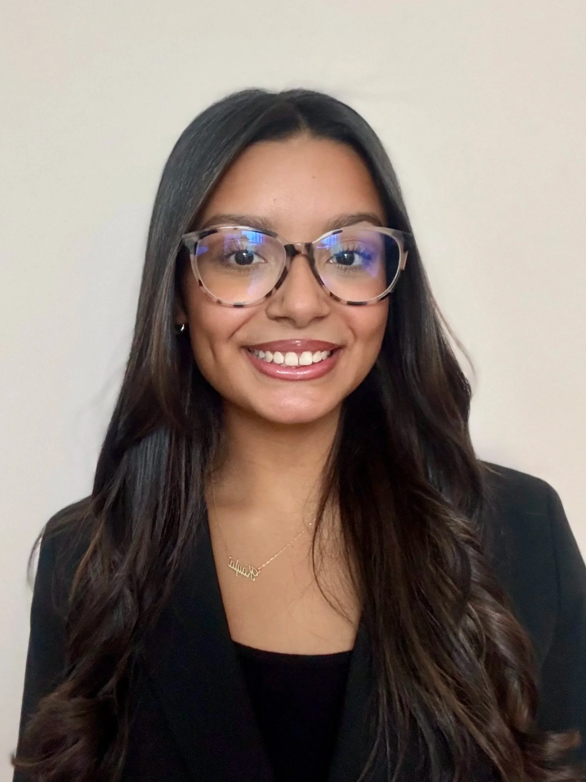 A smiling woman with long dark hair, glasses, a black blazer, and a gold necklace standing against a plain white background.