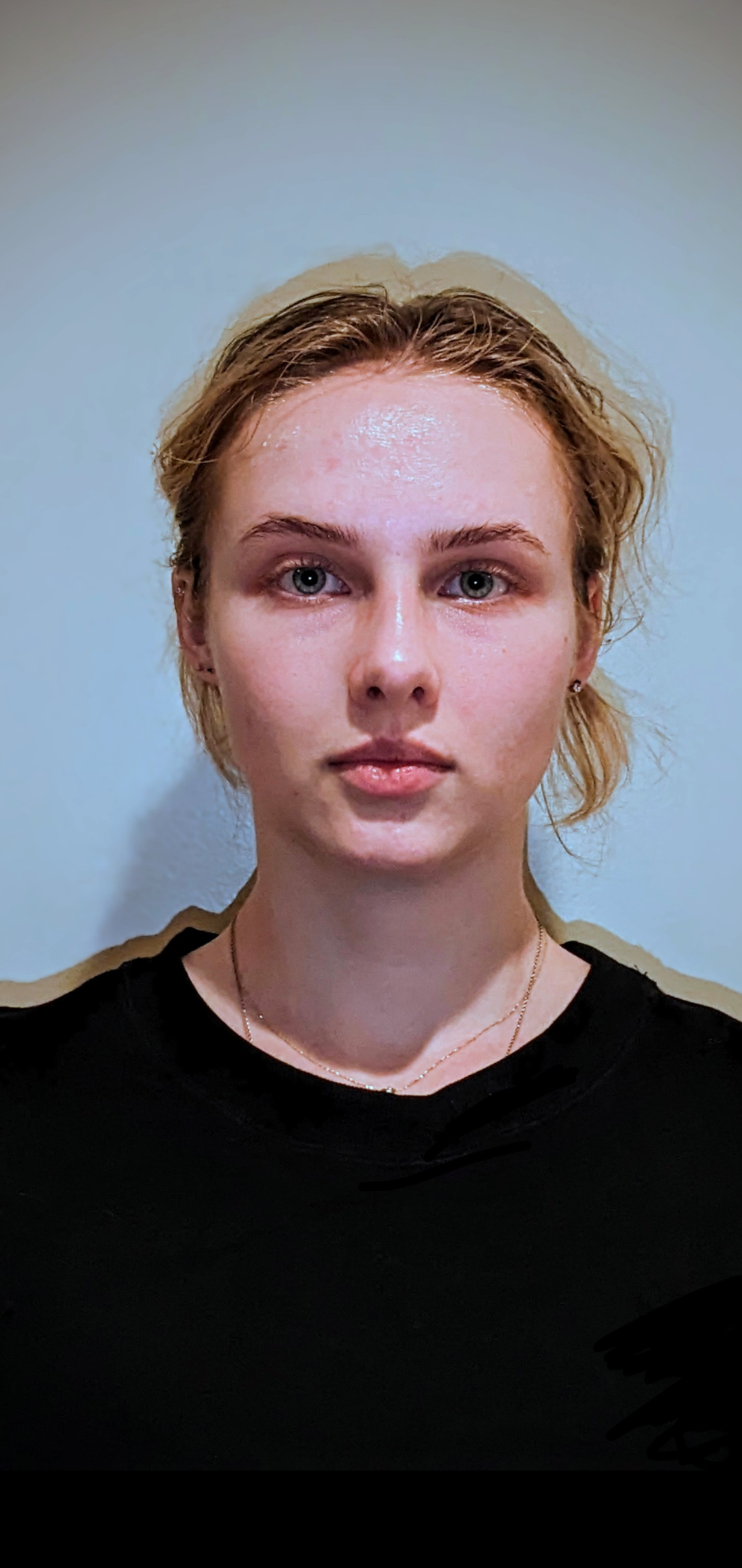 A portrait of a young woman with short, wavy blonde hair, blue eyes, wearing a black top and small earrings, standing against a light-colored wall.