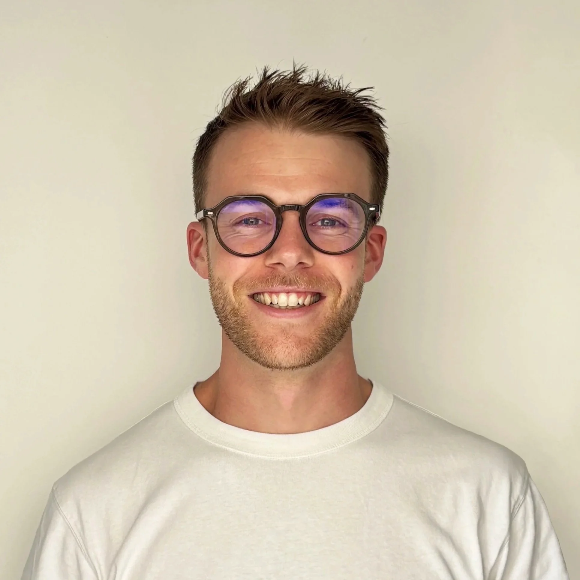 Portrait of a smiling young man with glasses, short brown hair, light beard, wearing a white t-shirt, standing against a plain light-colored background.
