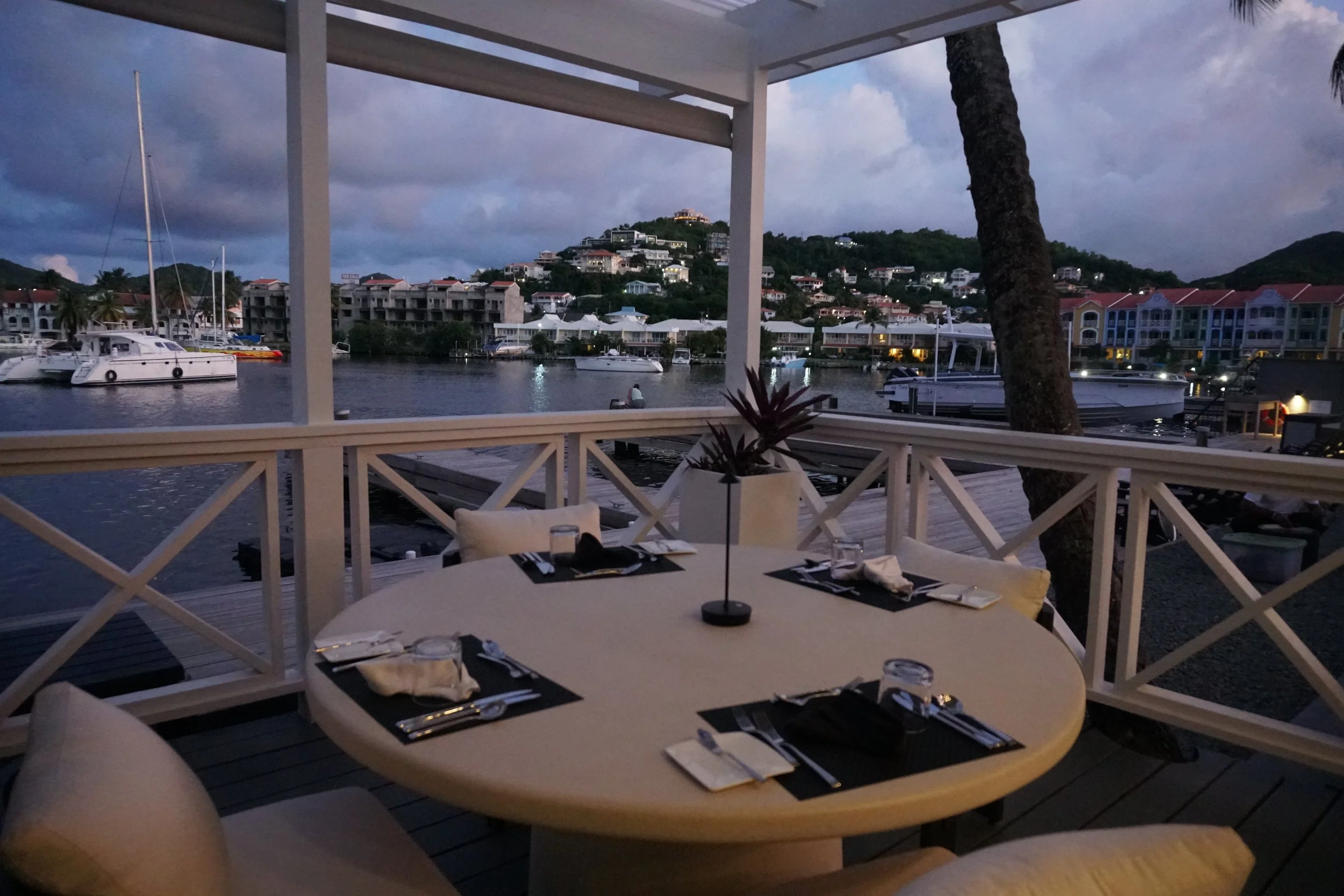 Dinner table set for four on a balcony overlooking a marina with boats, with a view of hillside houses at dusk.