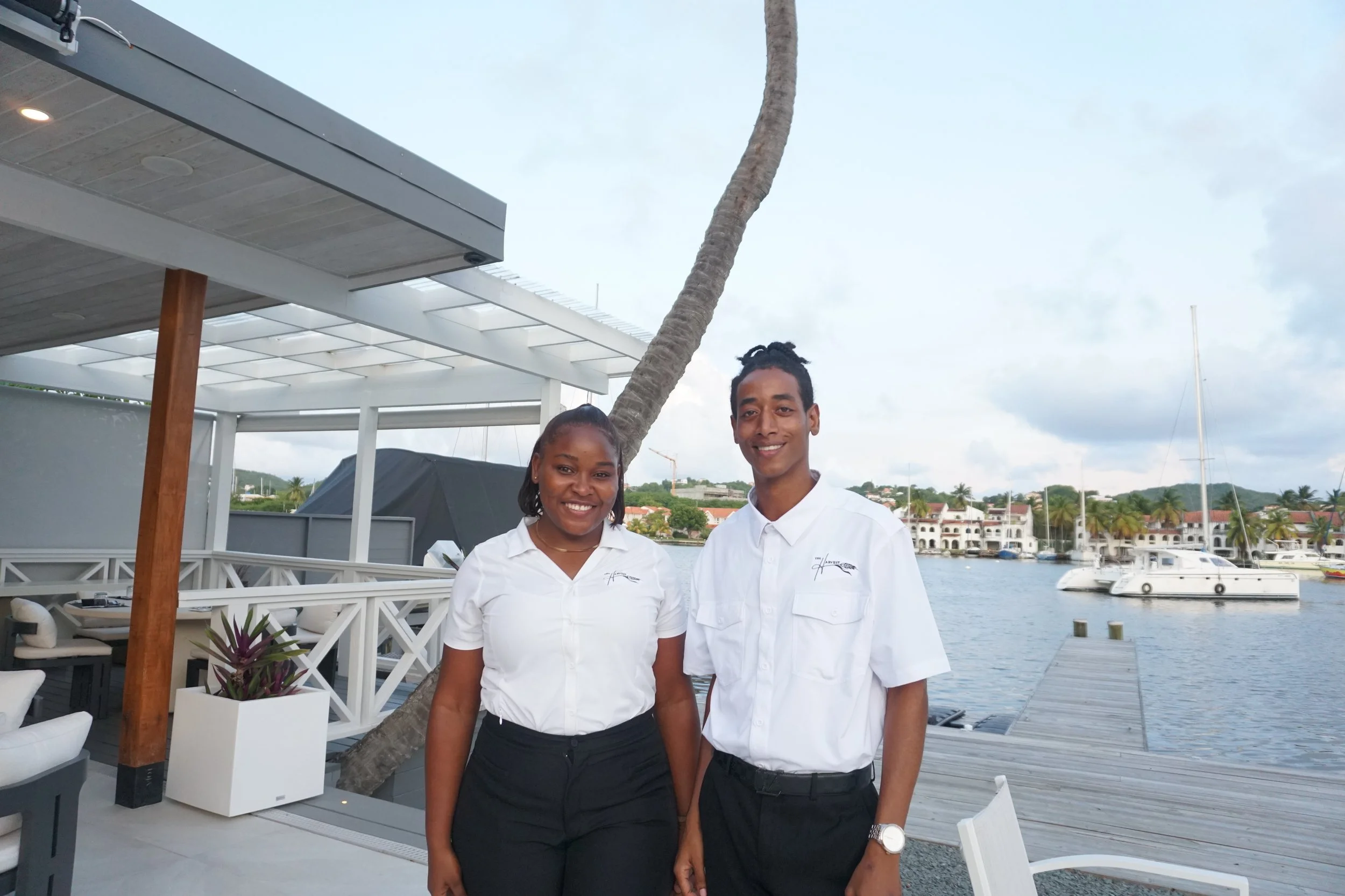 Two smiling people, a woman and a man, standing on a waterfront patio at The Harvest and Table Restaurant.