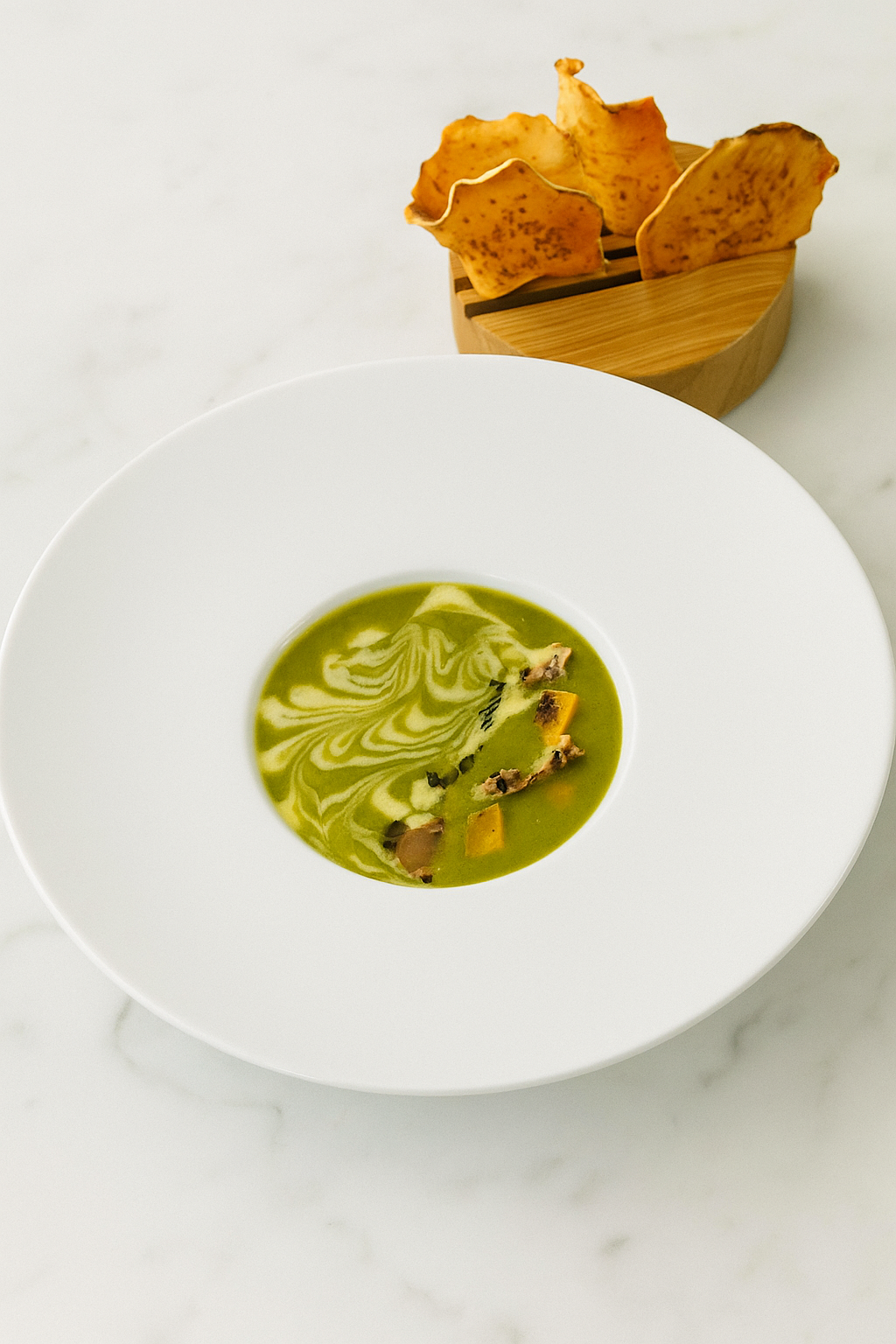 Green soup with swirled cream and garnishes served in a white bowl, with a wooden container holding dried fruit chips in the background.