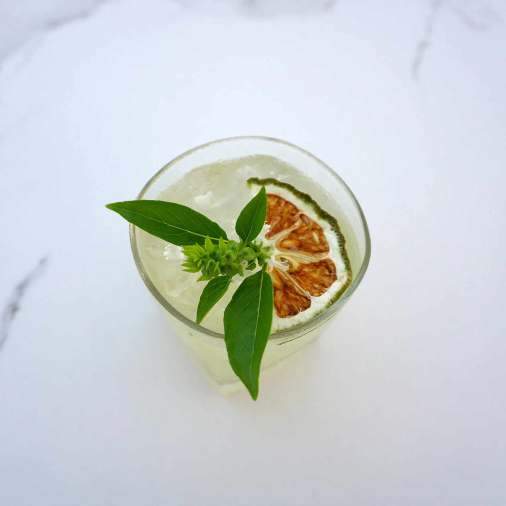 A clear glass of lemonade garnished with a dried lemon slice and fresh herbs, including basil leaves, on a white surface.