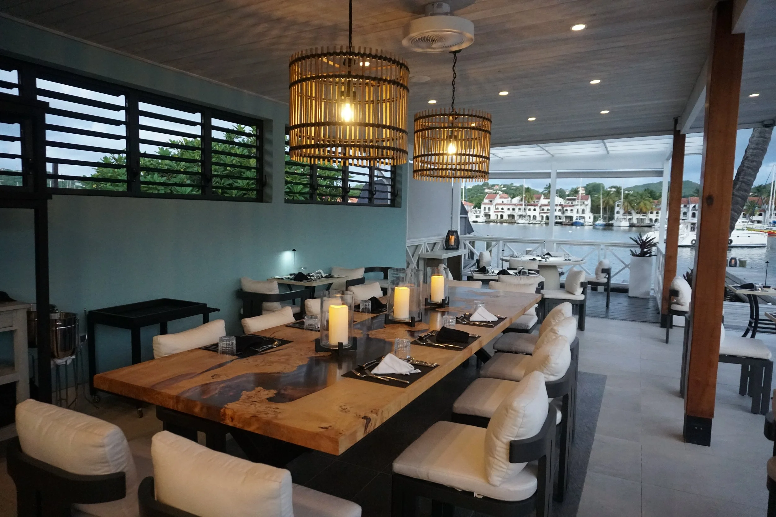 Elegant waterfront restaurant dining area with a long wooden table set with candles, candles, of candles, and tableware, overlooking a marina with sailboats and Mediterranean-style buildings.