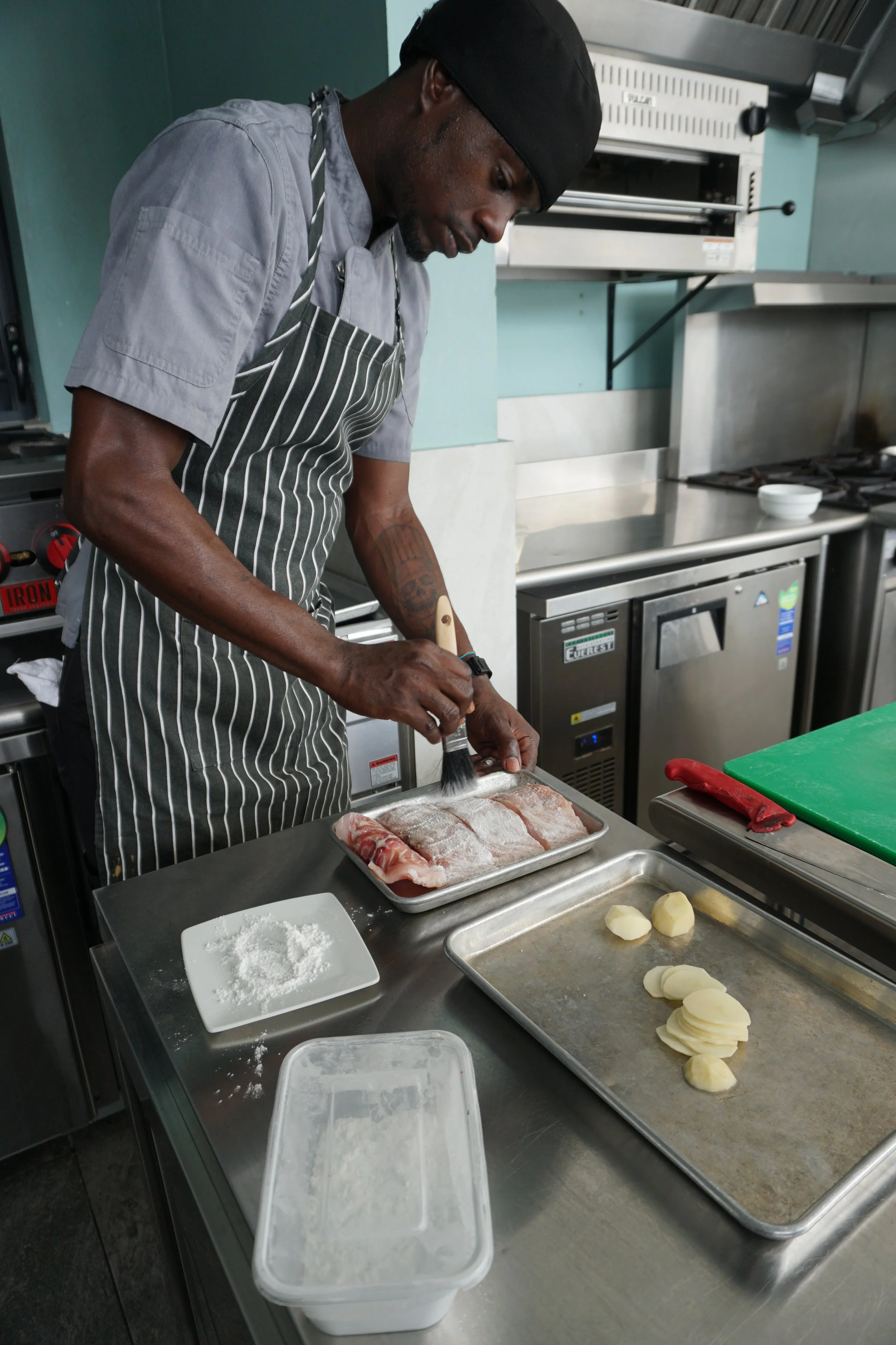 A man in a striped apron is seasoning raw fish fillets on a tray at The Harvest and Table Restaurant. 