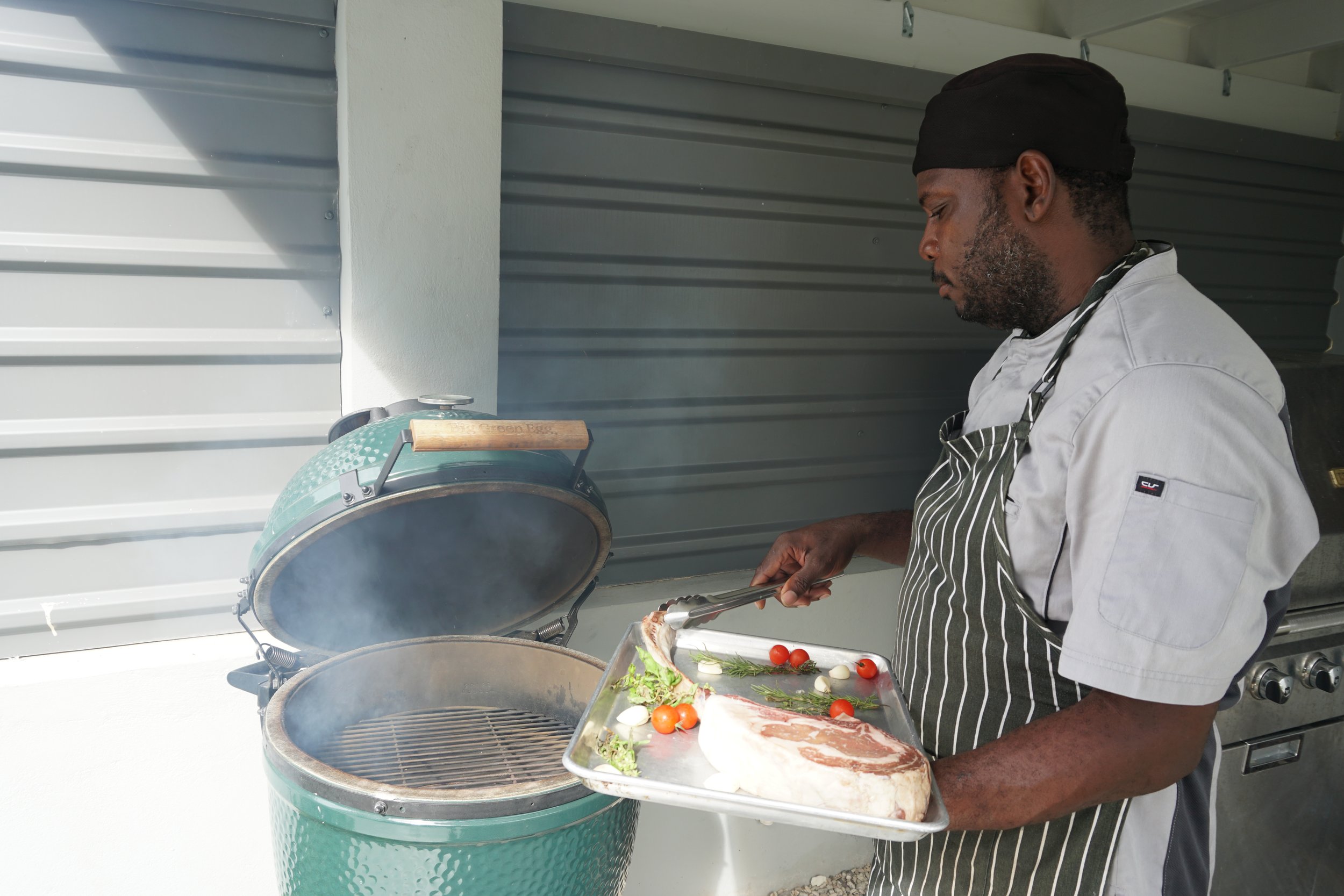 Man grilling a piece of meat on a green outdoor grill, holding a tray with vegetables and meat at The Harvest and Table Restaurant. 