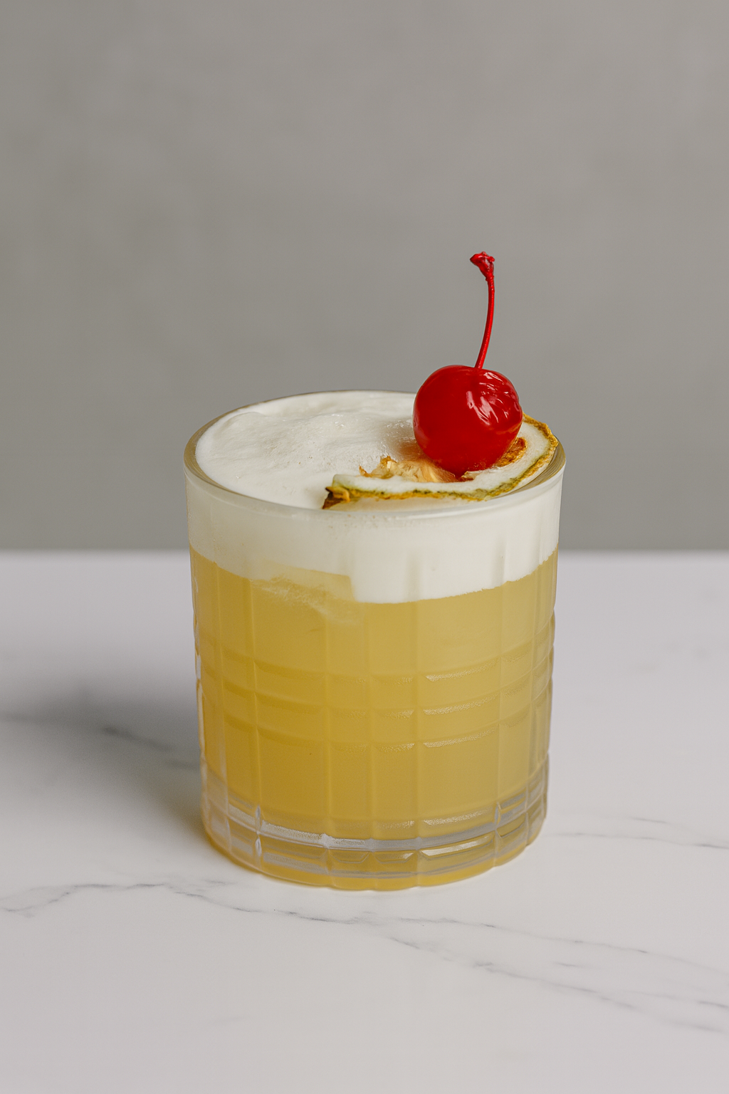 An amaretto sour in a textured glass topped with white foam, garnished with a maraschino cherry and a lemon slice.