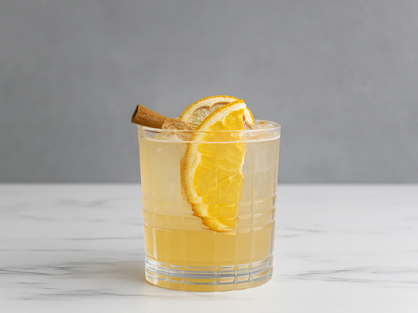A clear glass with a yellow beverage, garnished with lemon slices, a cinnamon stick, and ice cubes on a white marble surface against a gray background.