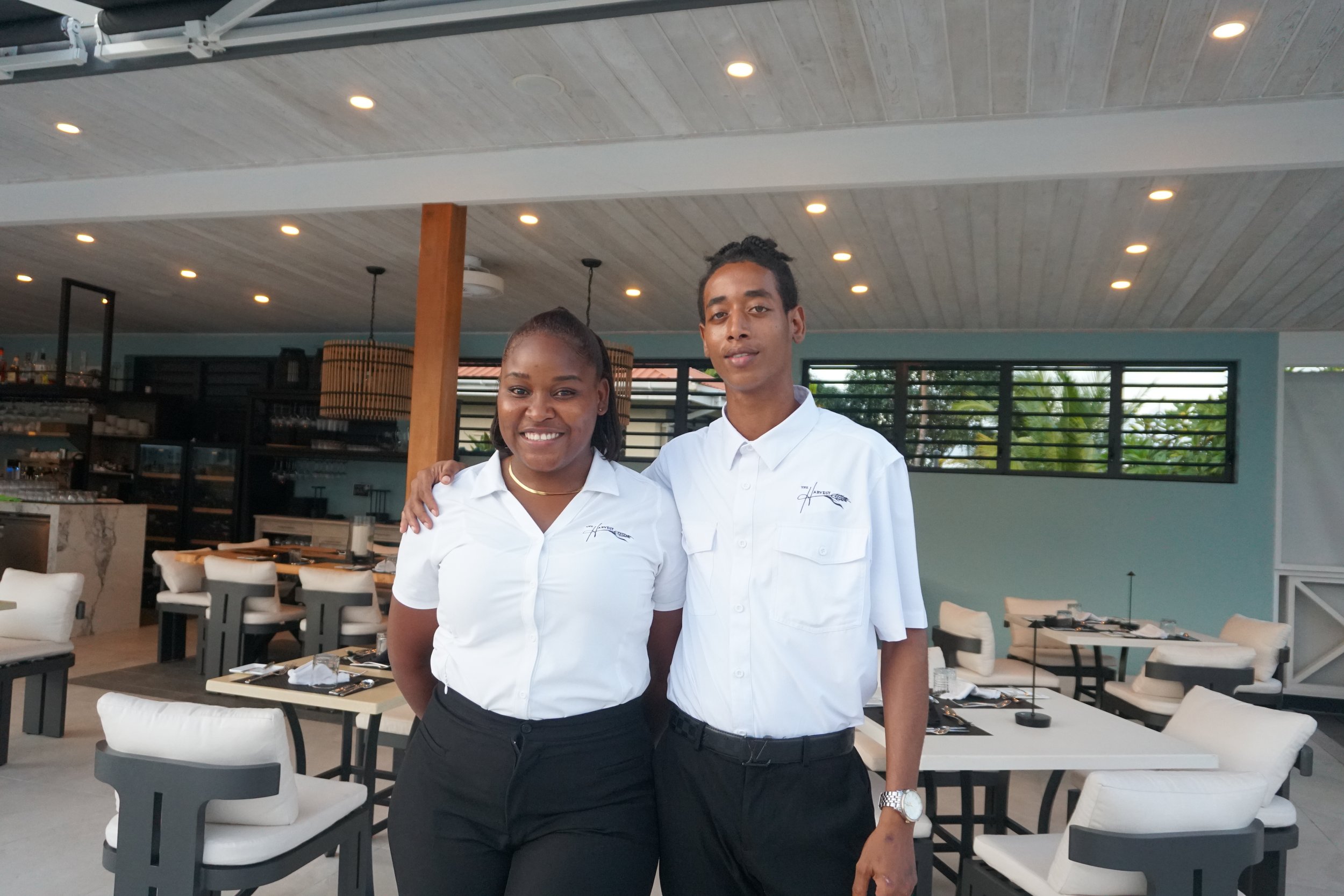 Two restaurant staff members, a woman and a man, wearing white uniforms, standing together at The Harvest and Table Restaurant. 