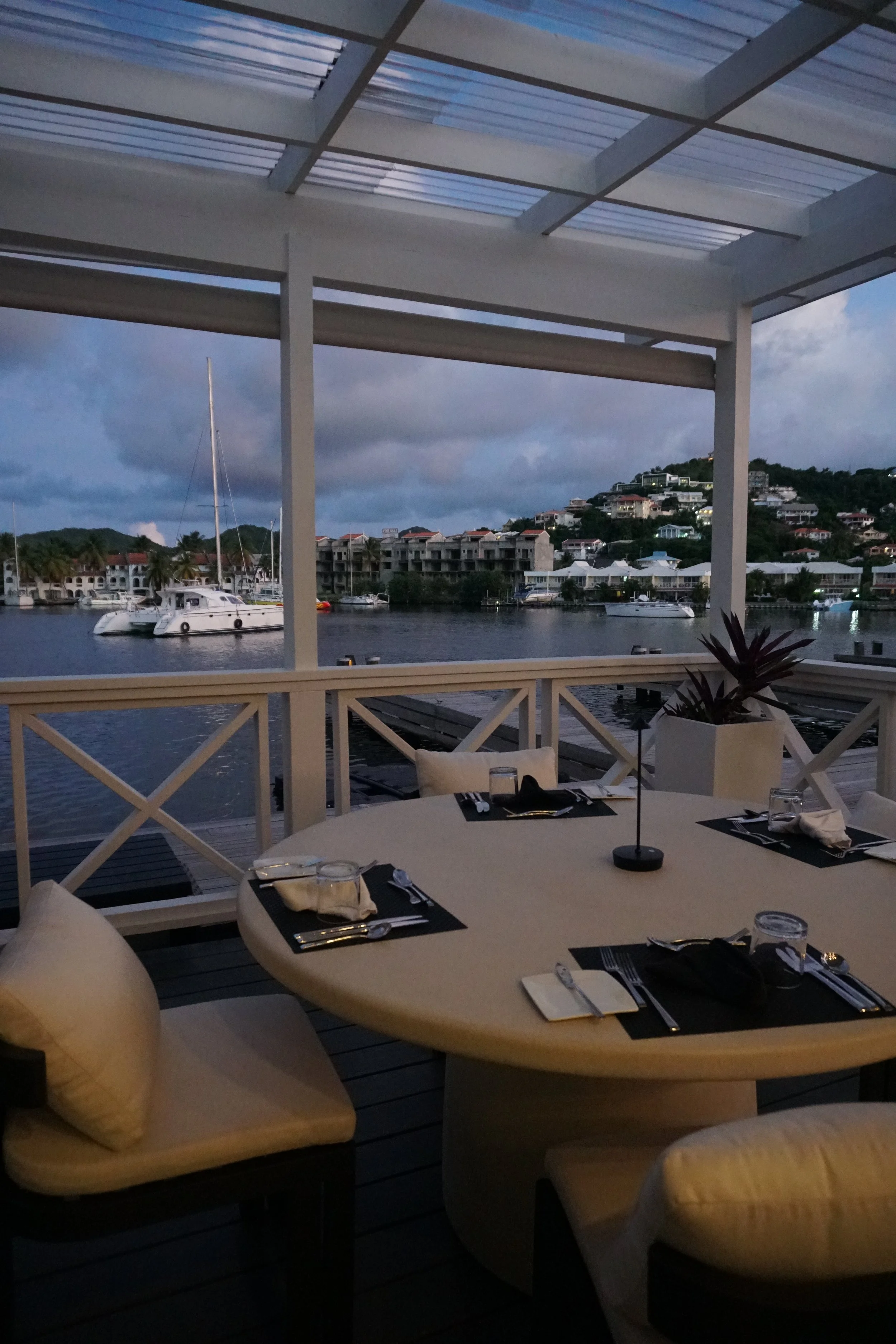 A table set for dining on a covered outdoor patio overlooking a marina with boats and houses on a hillside.