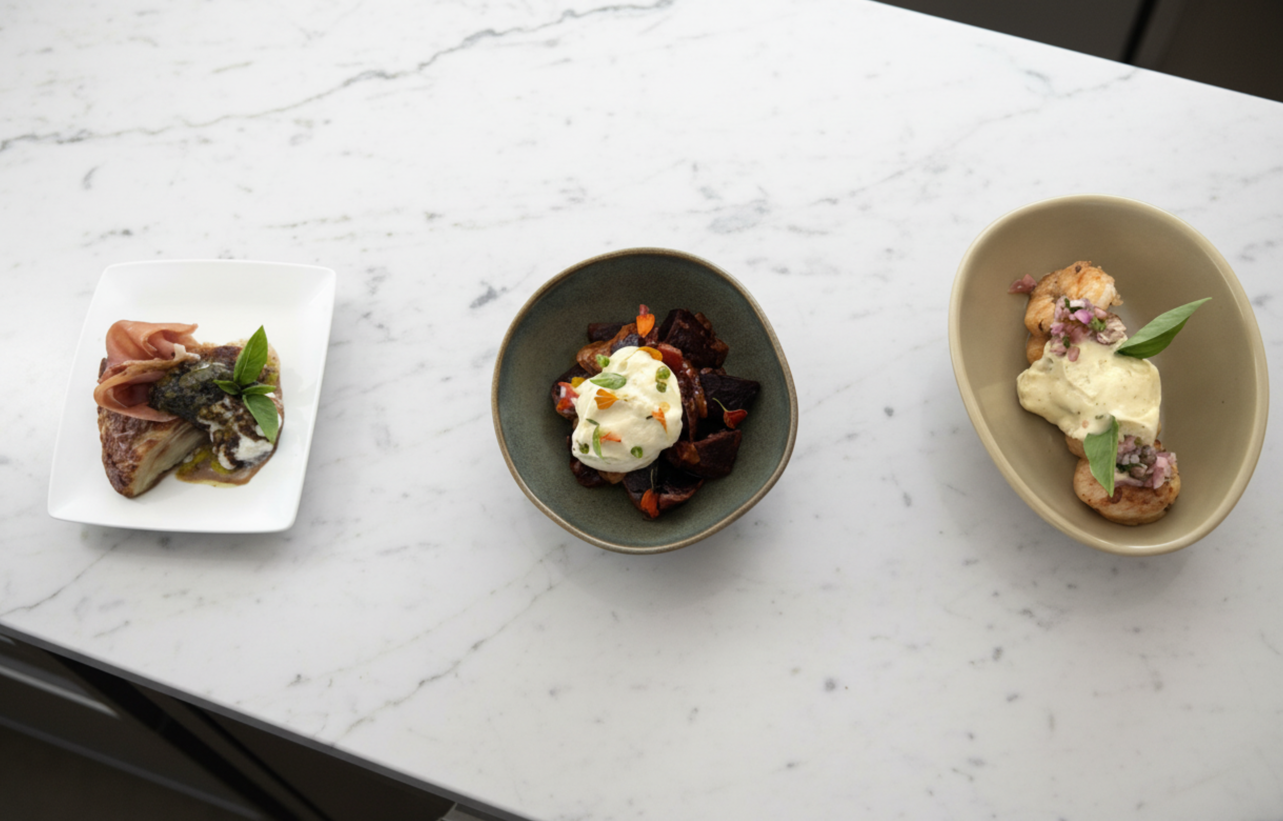 Three plates of gourmet food on a white marble surface, from left to right: a serving of meat with sauce, a bowl of roasted vegetables topped with cream, and a crostini with creamy topping garnished with herbs and edible flowers.