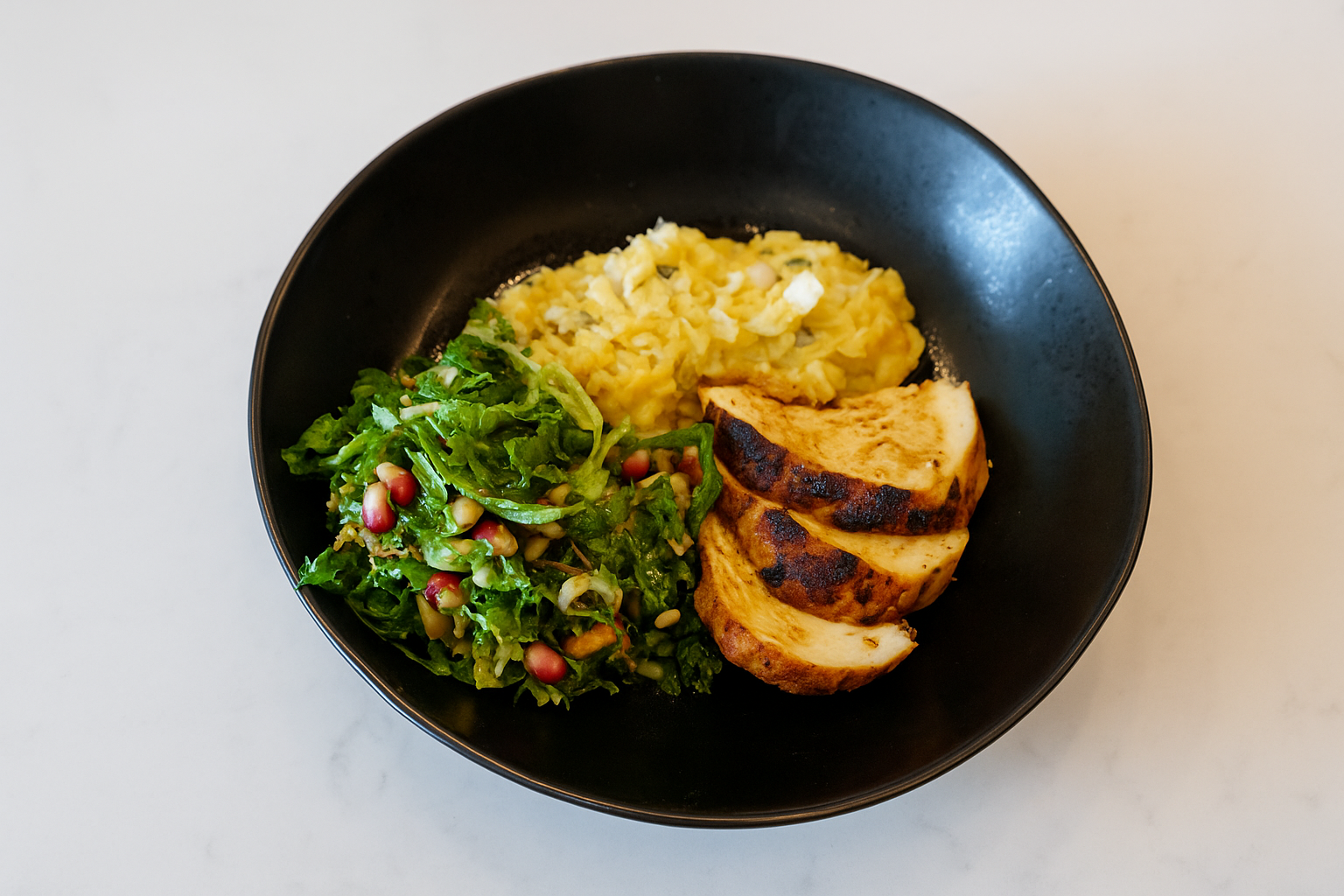 Black bowl containing scrambled eggs, grilled chicken slices, mashed potatoes, and a green salad with pomegranate seeds.