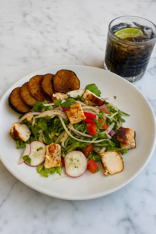 A white plate with a salad containing grilled chicken, sliced radishes, chopped tomatoes, onions, and leafy greens, served with fried plantains on the side. A glass of dark soda with lime slices is in the background on a white marble surface.