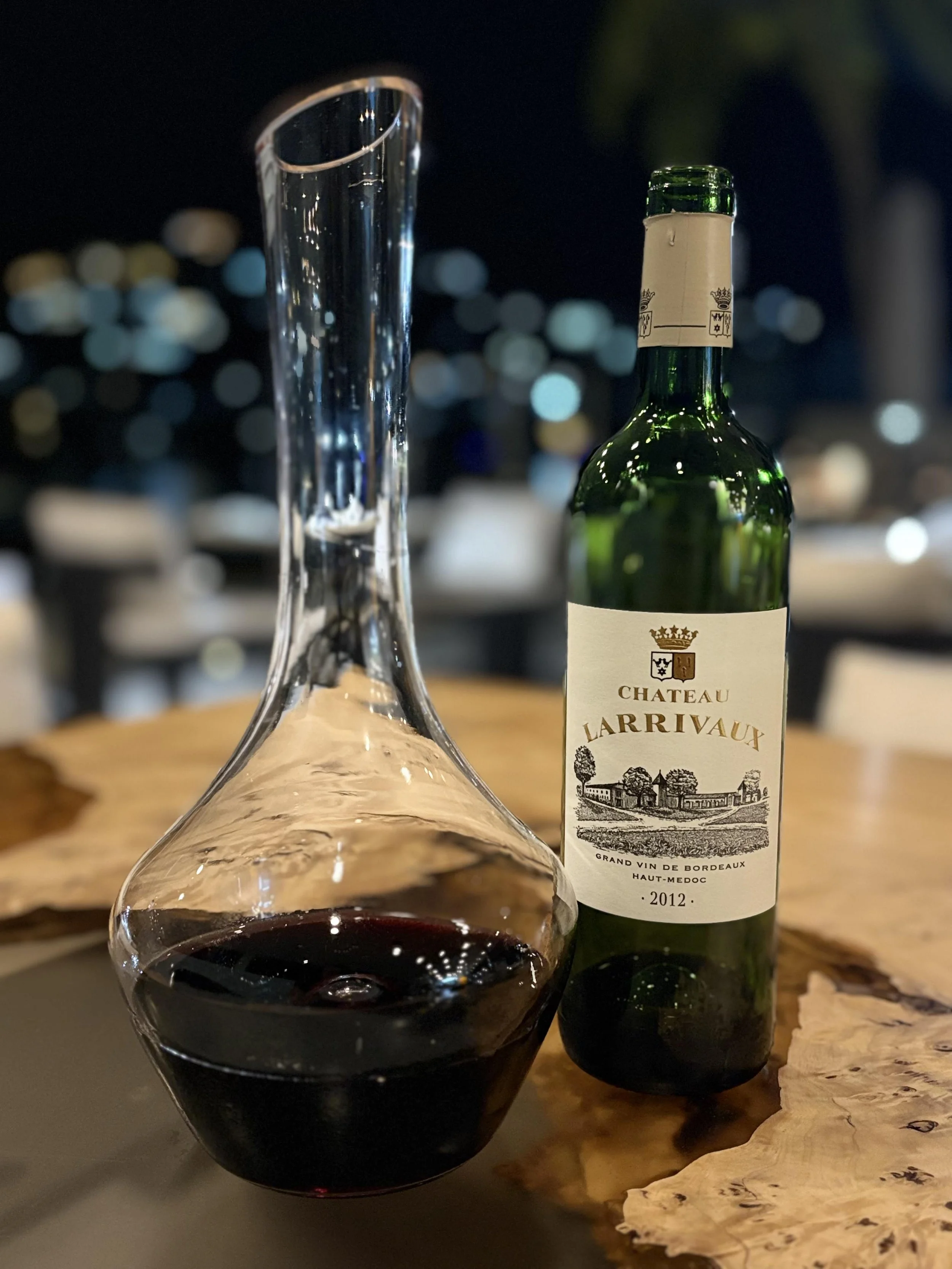 A glass decanter filled with red wine placed on a wooden table, with a bottle of Château Larrivaux Bordeaux wine from 2012 next to it, against a blurred nighttime cityscape background.