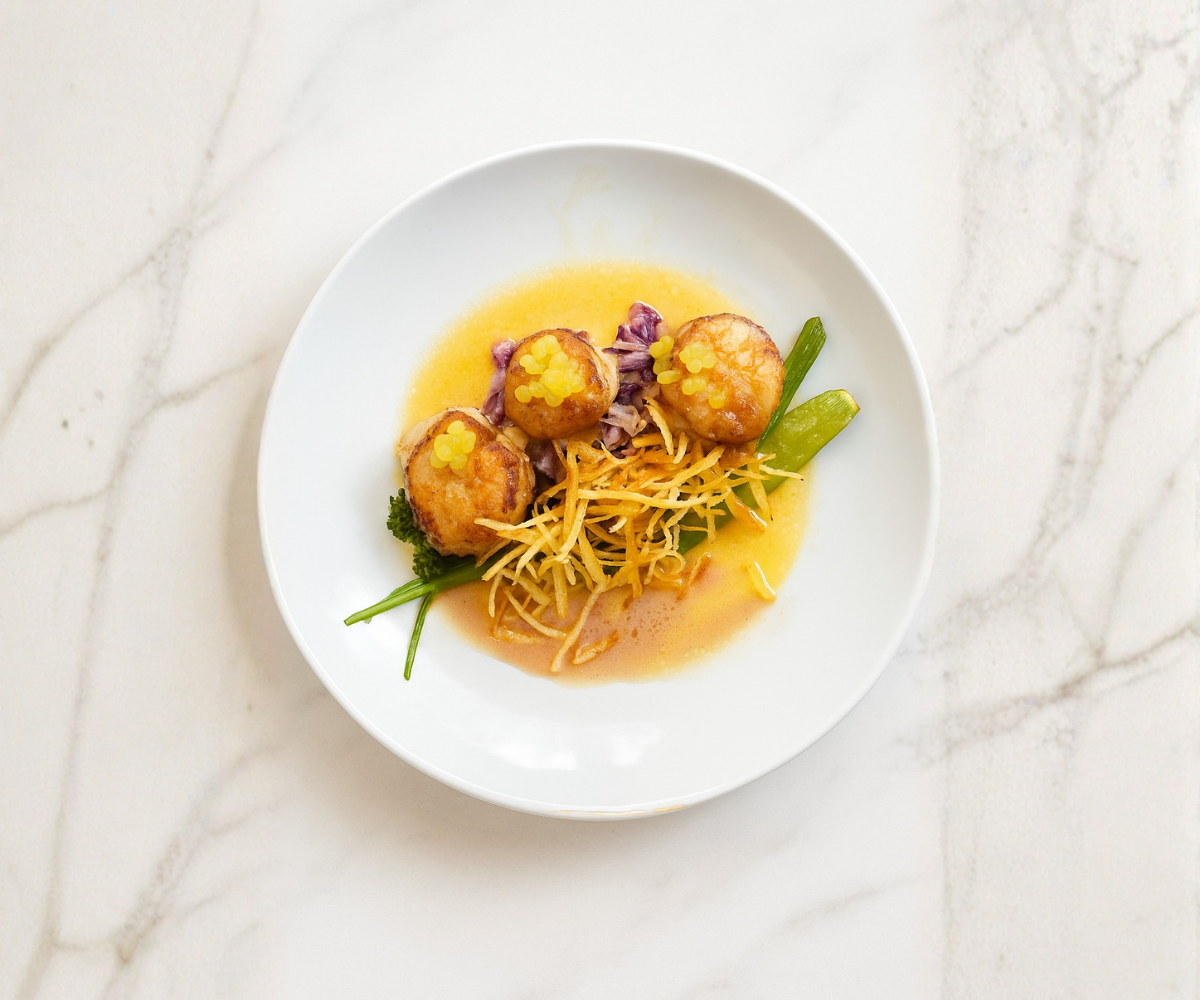 Plate with three browned meatballs topped with yellow sauce pearls, shredded cheese, green beans, and greens, on a white plate on a marble surface.