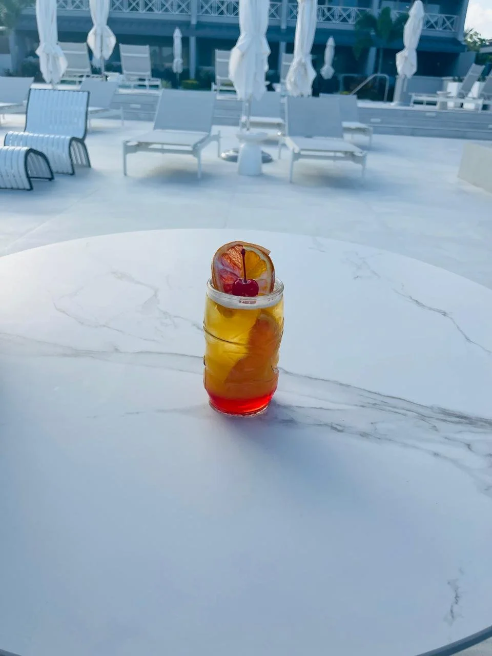 A colorful layered cocktail in a glass jar with a cherry and citrus slice garnish, placed on a white marble table against outdoor poolside lounge chairs and umbrellas.