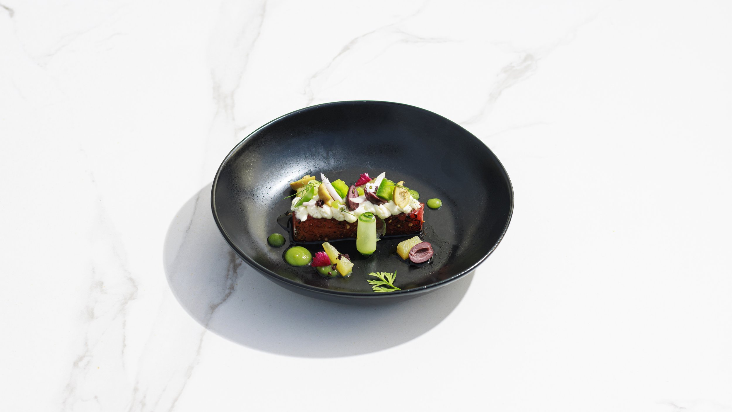 A black bowl with a gourmet dish consisting of a rectangular piece of meat topped with cream, diced vegetables, herbs, and sauce, garnished with colorful edible decorations and microgreens, on a white marble surface.