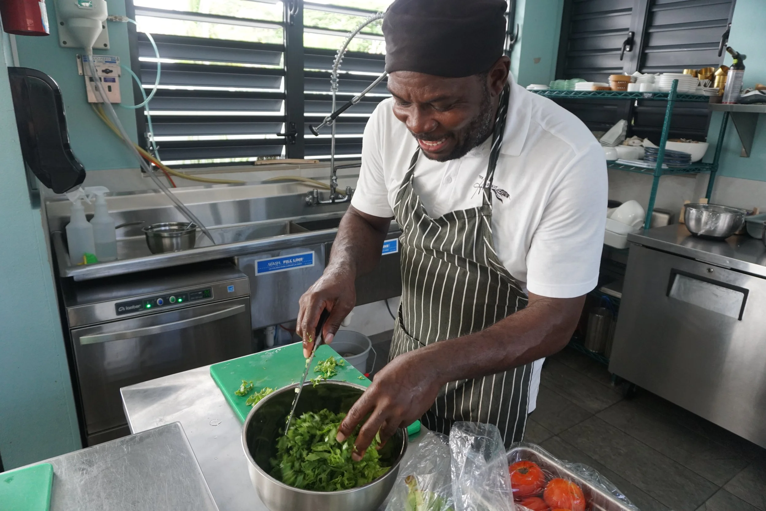 A man wearing a black head covering, white shirt, and striped apron mixing chopped leafy greens in a stainless steel bowl at The Harvest and Table Restaurant.