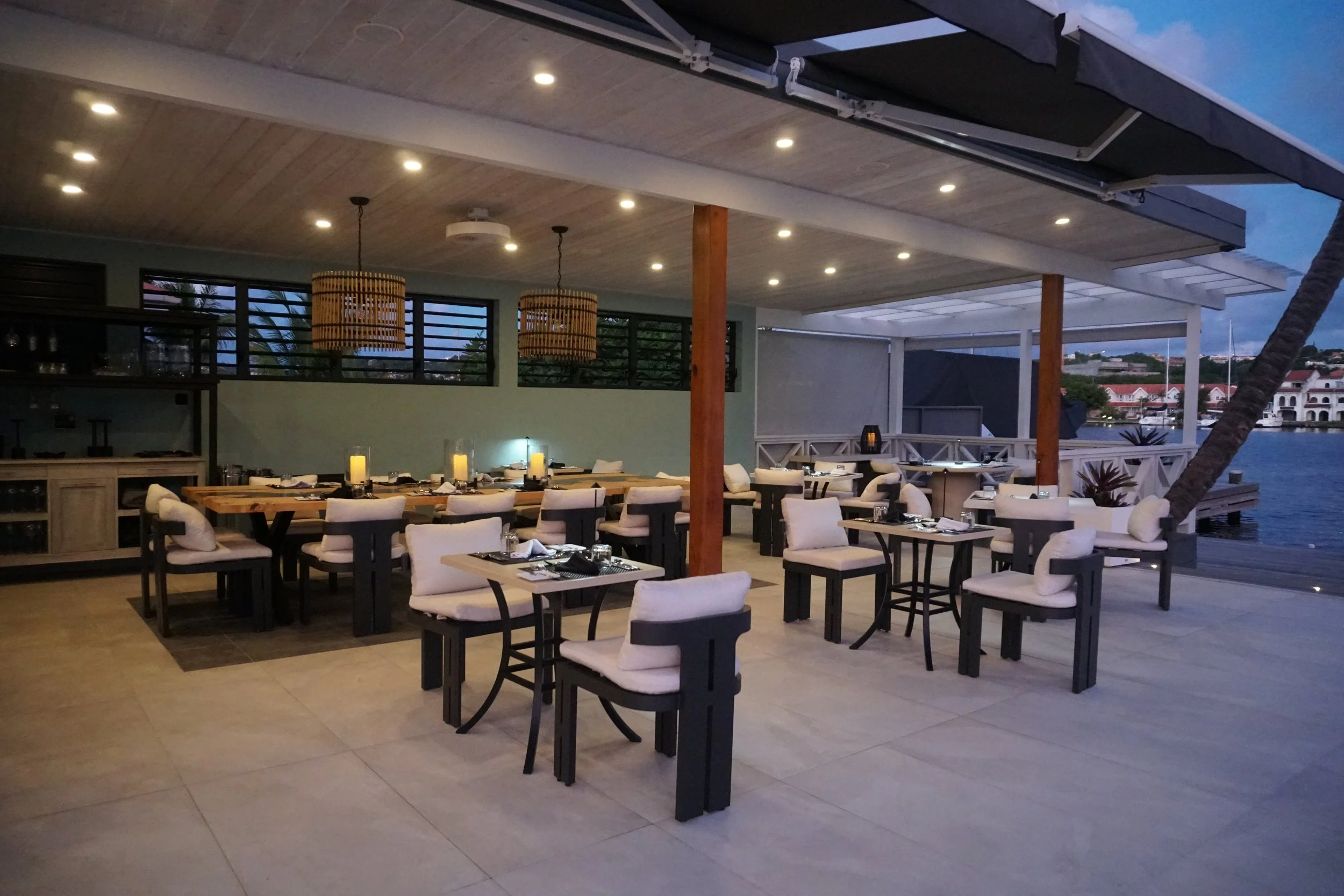 Dining area at a waterfront restaurant during twilight with tables set with black plates, cutlery, and candles, featuring a covered patio with hanging lights and a view of water and buildings in the distance.