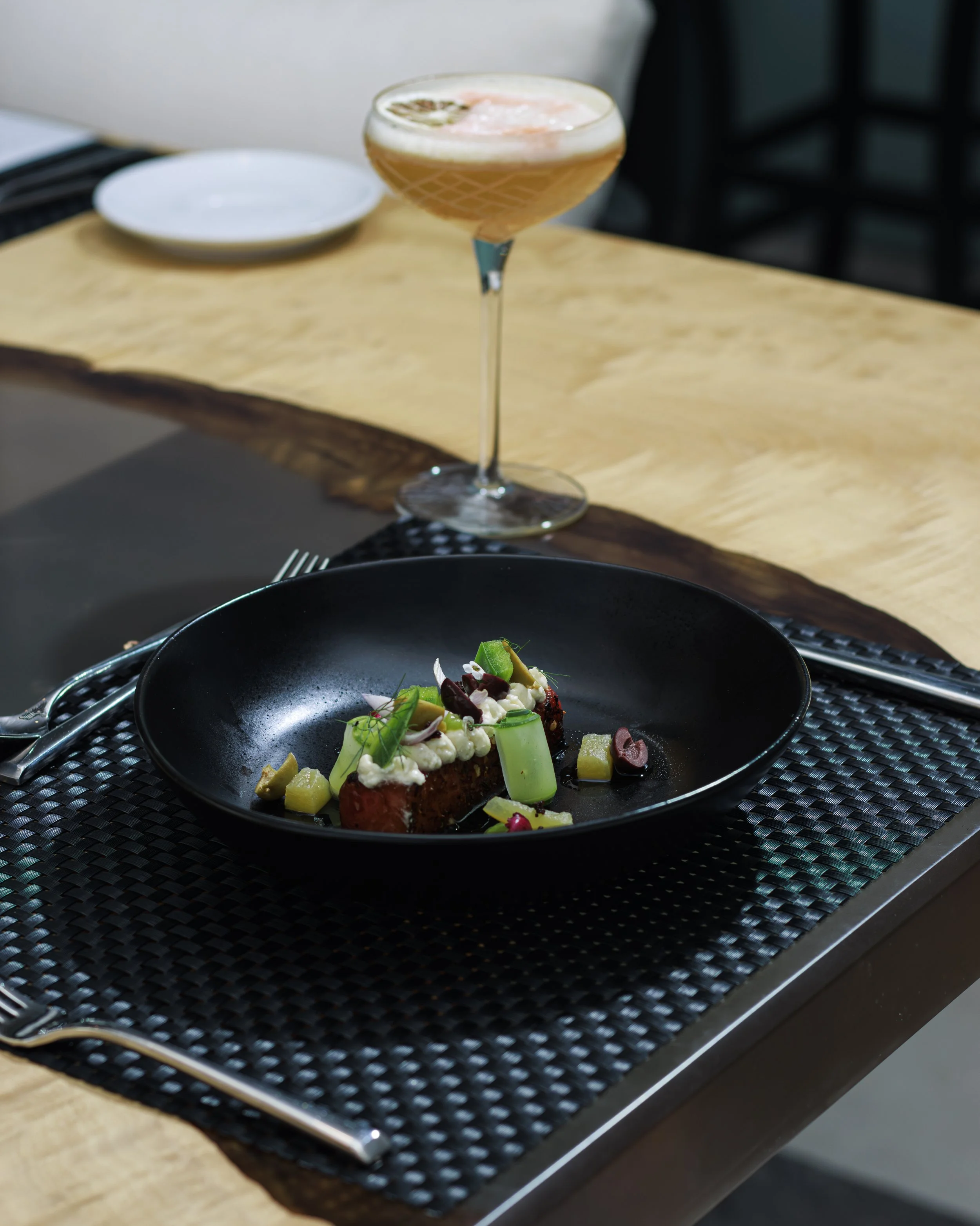 A gourmet plate with a piece of meat topped with white sauce, garnished with green herbs and vegetables, on a black plate. Behind it, a cocktail with a lemon slice, served in a tall glass.