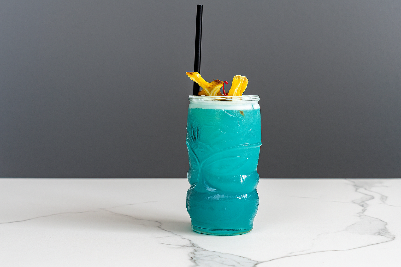 A tall, wavy glass with a bright blue cocktail, garnished with pineapple slices, a cherry, and a black straw, placed on a white marble surface with a plain gray background.